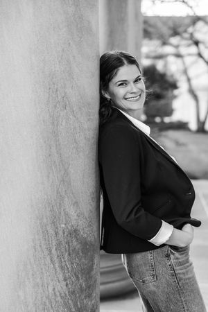 Black and white photo of a smiling young woman leaning against a stone column outdoors, wearing a blazer and jeans.