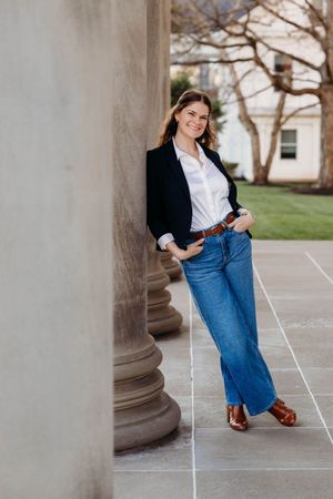 Young woman in blazer and jeans leaning against stone column outdoors, smiling confidently in a campus setting.