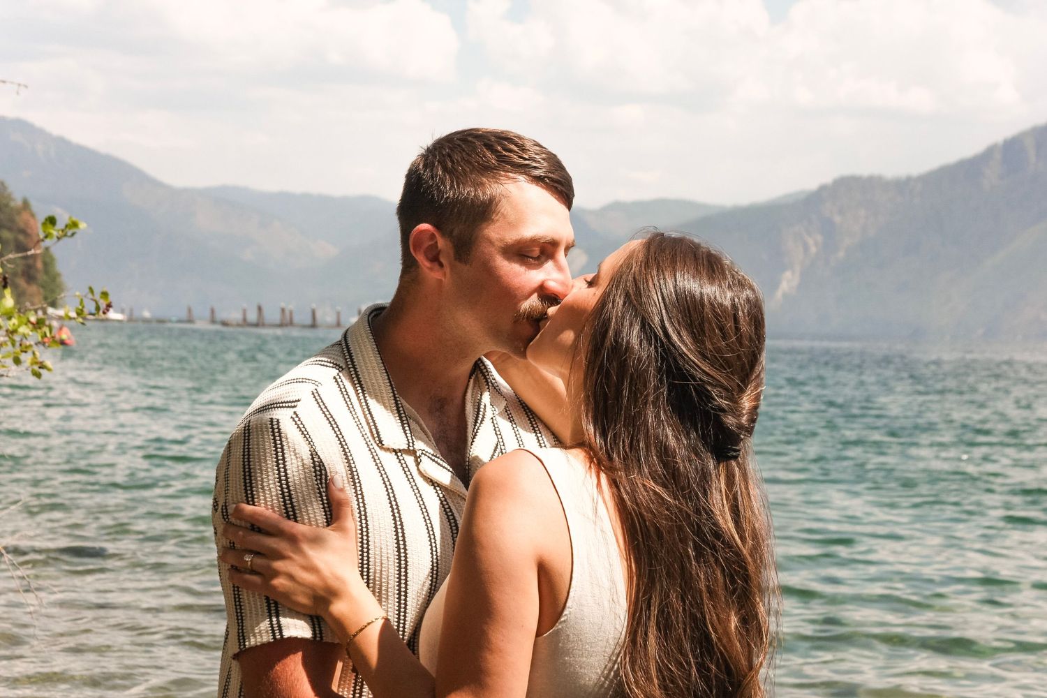 Couple kissing romantically by a scenic mountain lake on a sunny day.