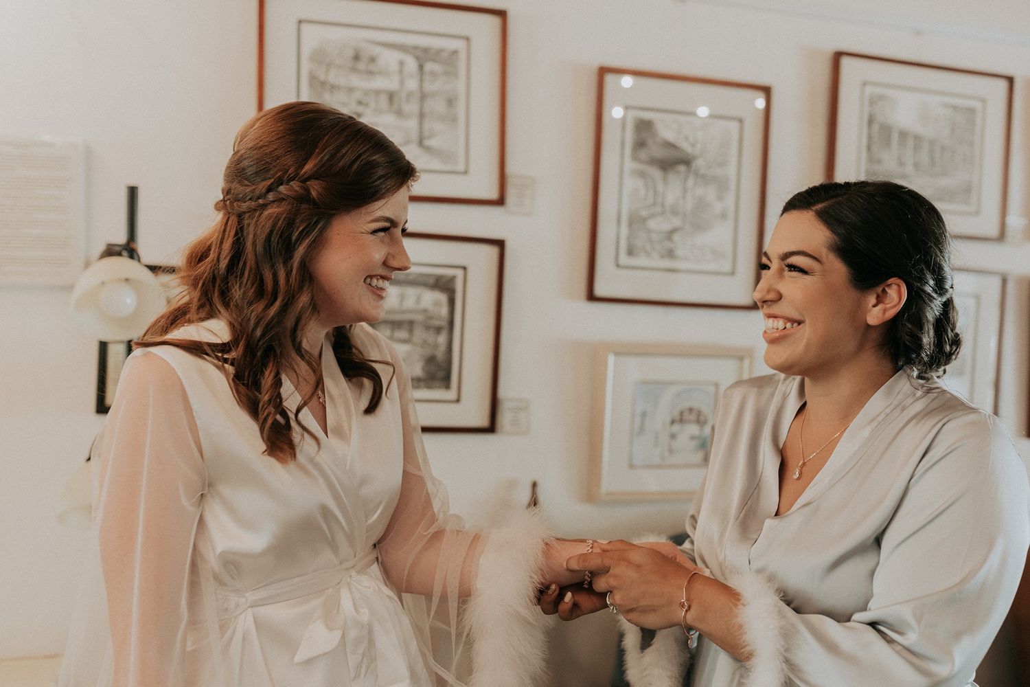 Two bridesmaids in satin robes laughing and holding hands during wedding preparations indoors.