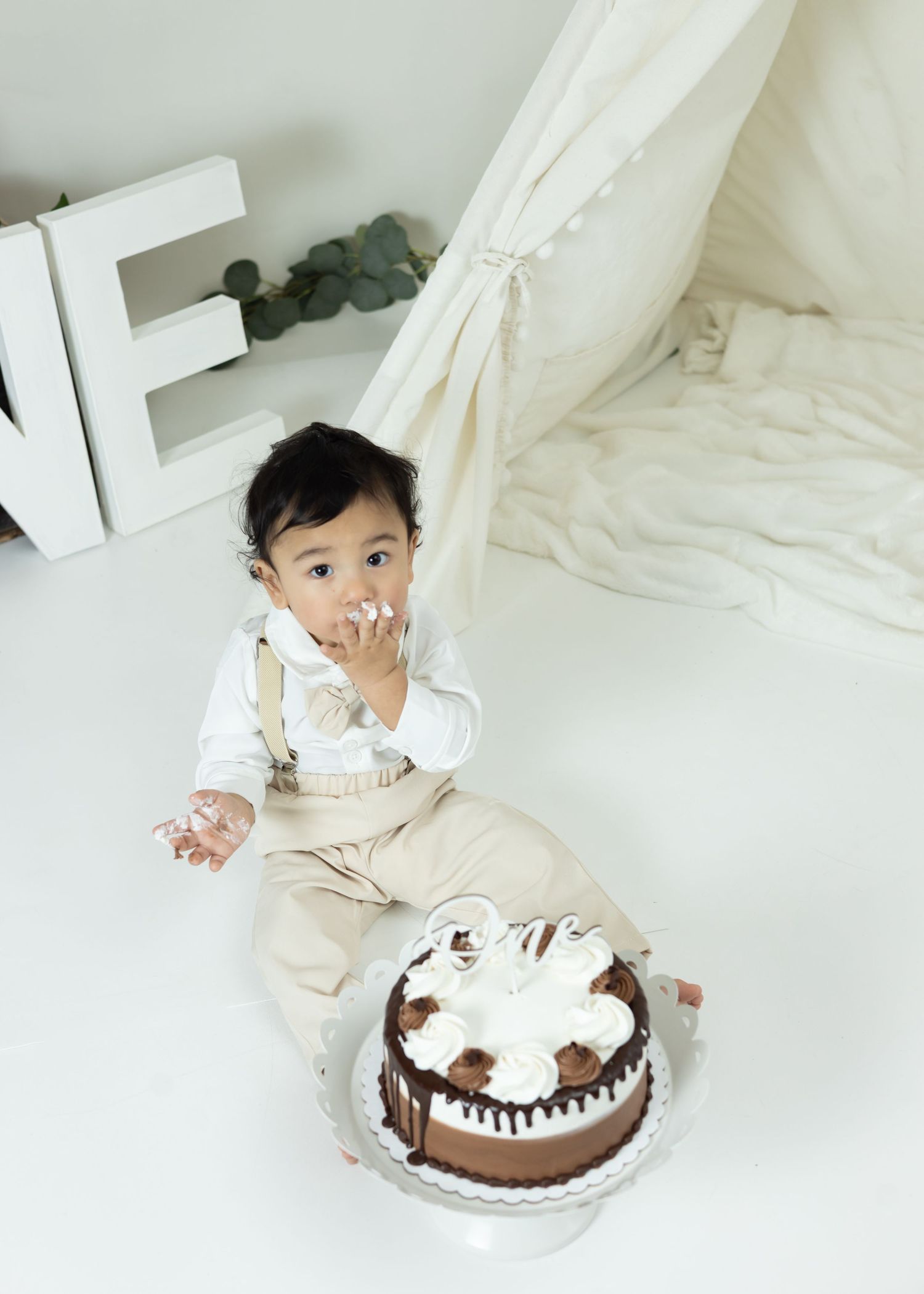A small child in cream-colored clothing plays with a chocolate birthday cake on a white backdrop.