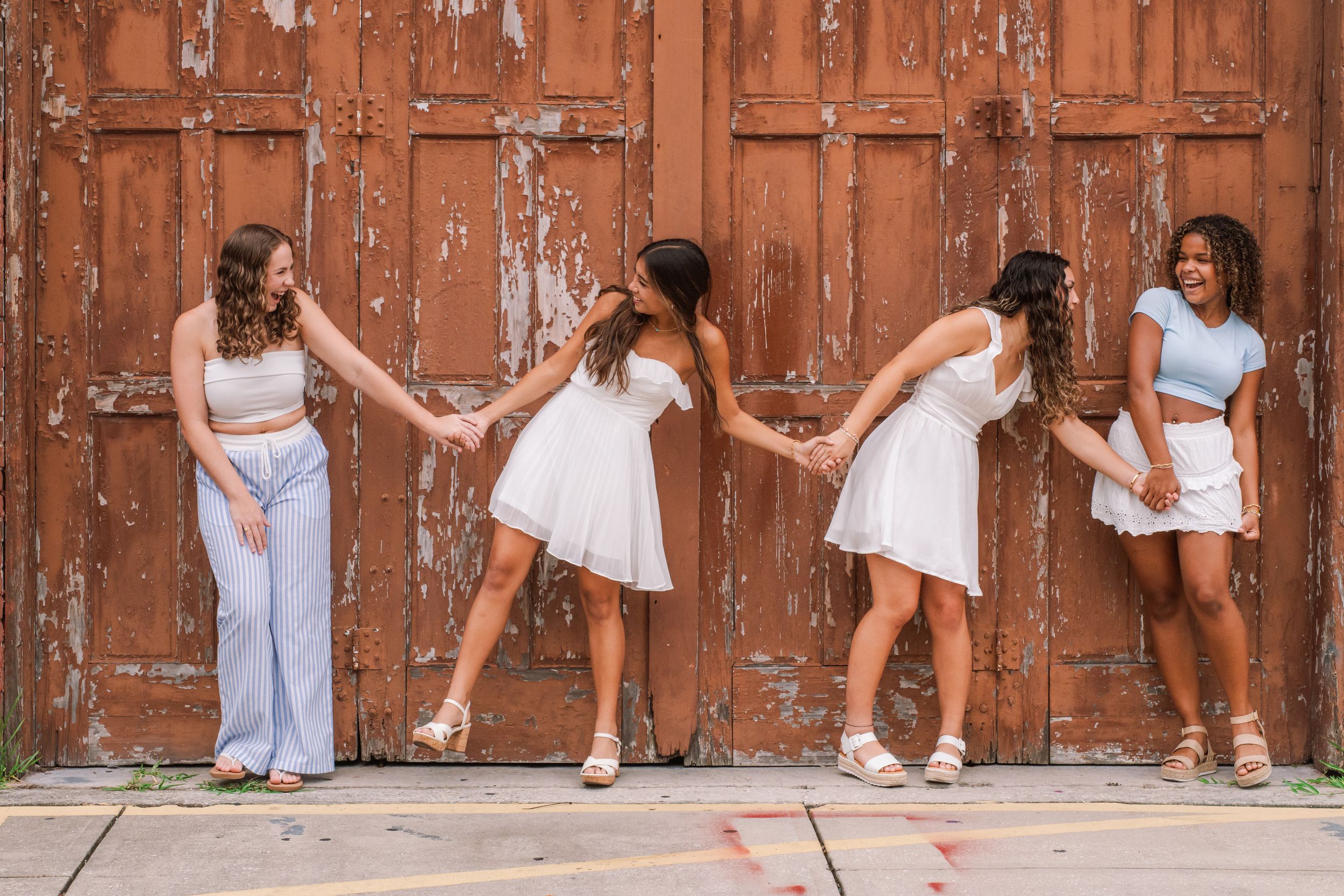 Four young women in white and pastel outfits hold hands in a chain pose against a rustic wooden door.
