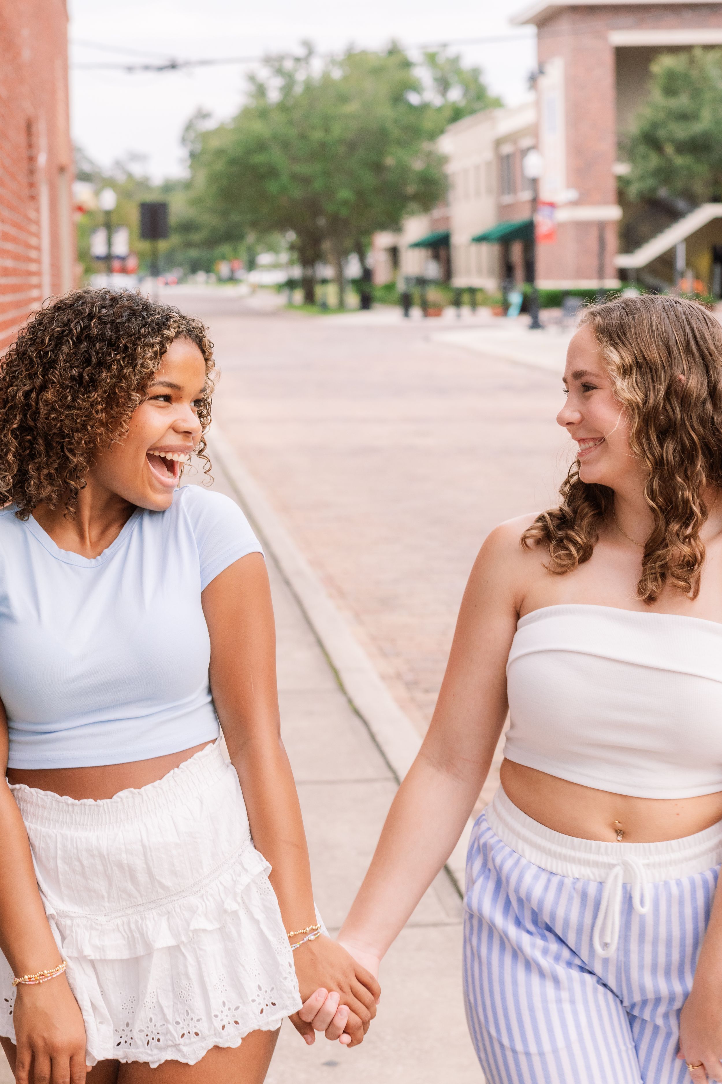 Two young women holding hands and laughing together while walking on a sunny urban sidewalk.