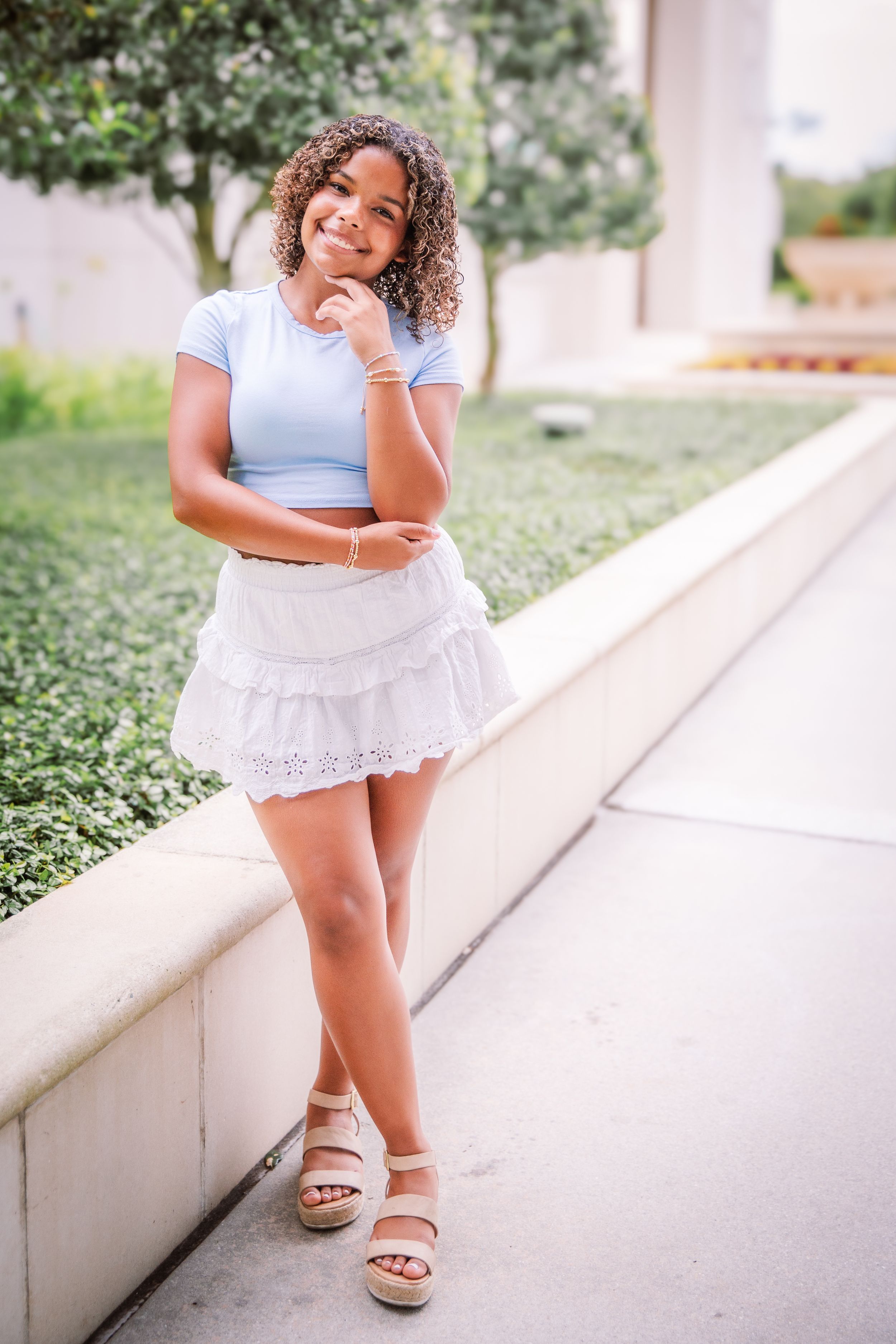 A person in a light blue top and white lace skirt poses playfully on white stone steps in an outdoor setting.