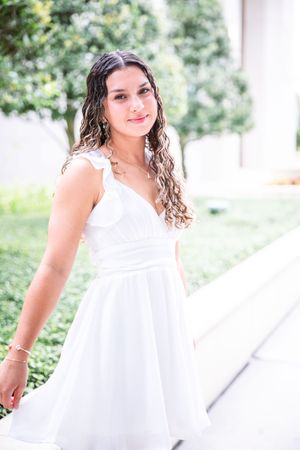 A person in a flowing white dress with curly hair poses outdoors against a green garden background.