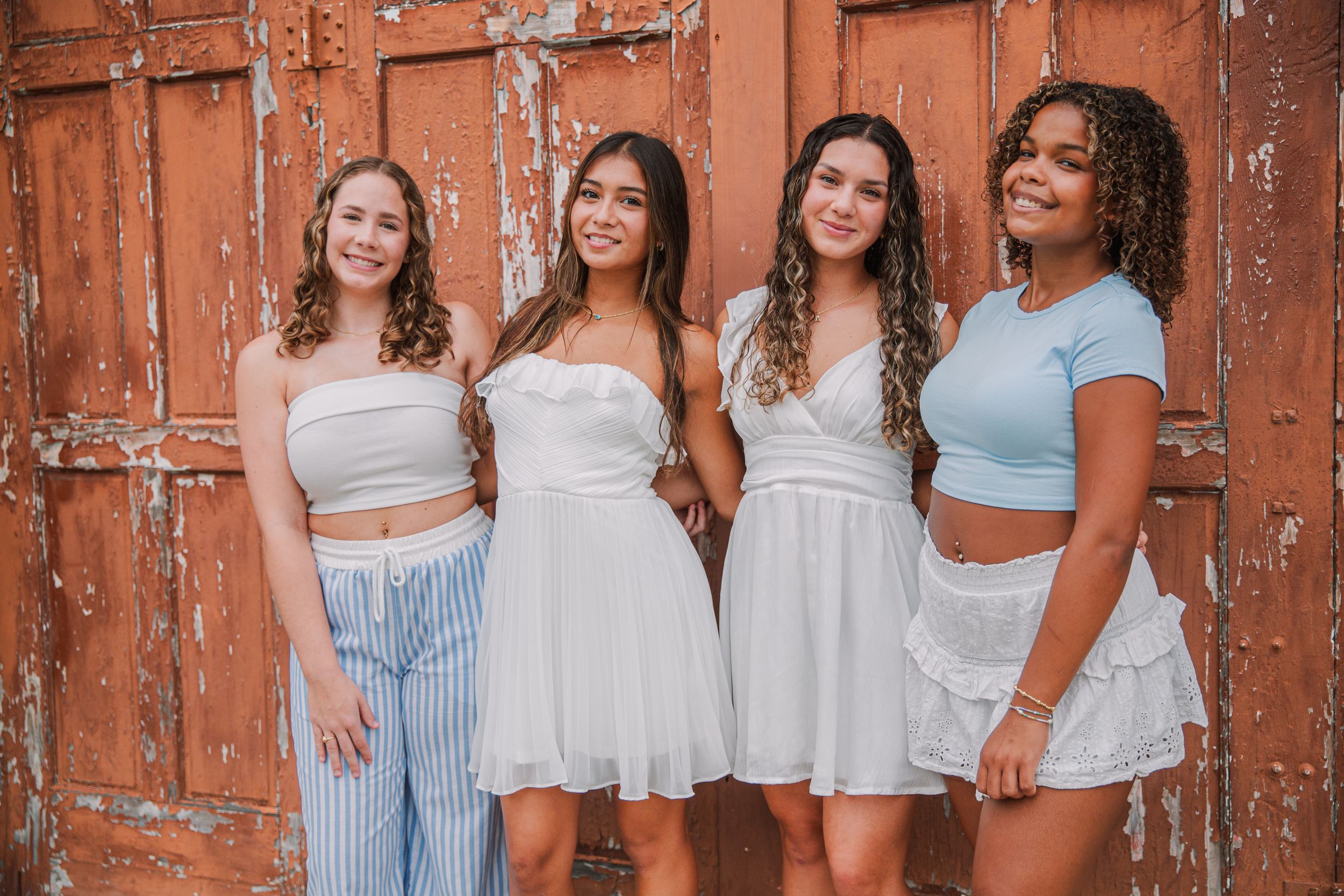 Four young women in white and pastel outfits posing together in front of a rustic red wooden door.