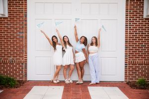 Four young women in white outfits pose joyfully in front of a brick building holding small pennant flags.
