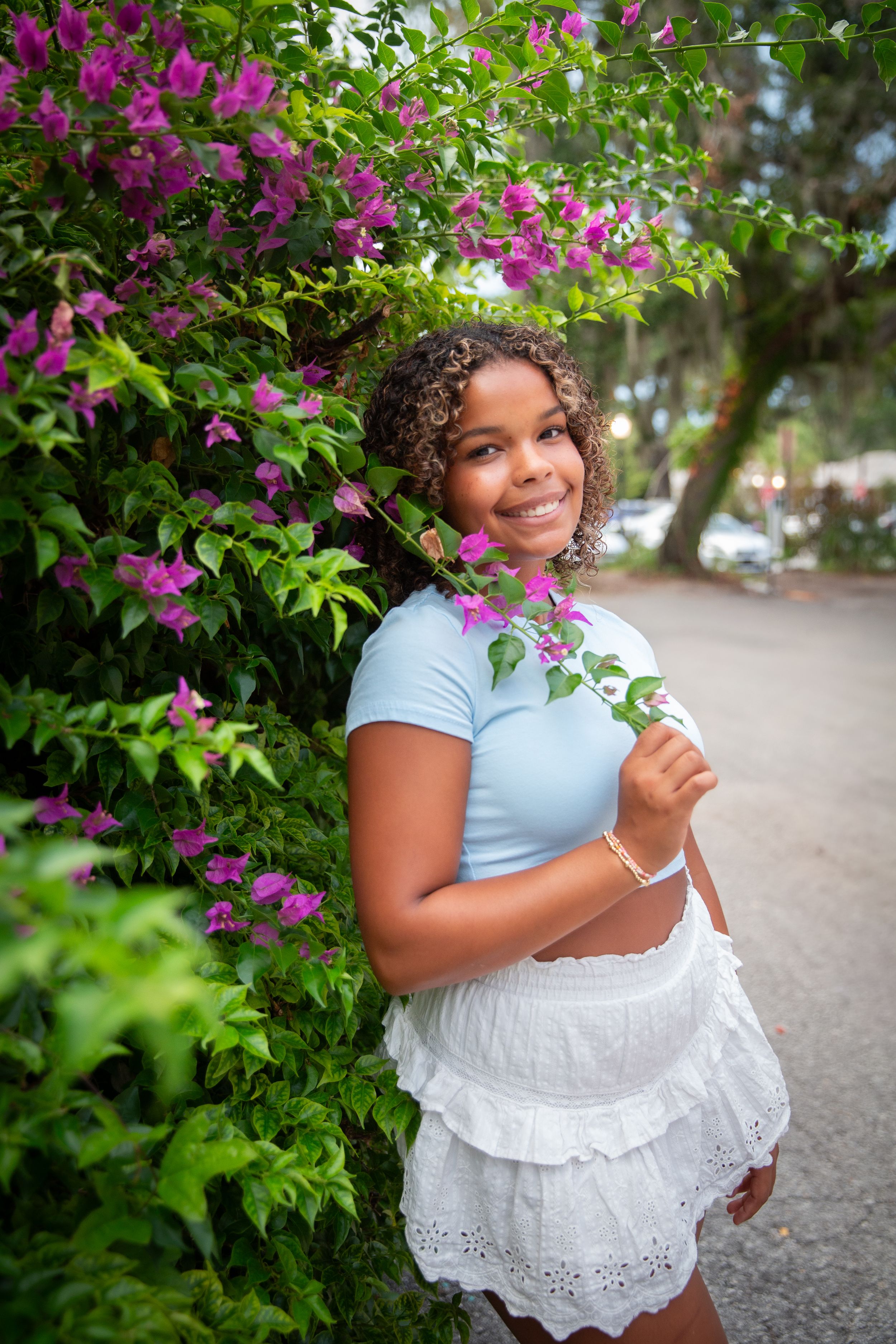 A series of photos showing someone in a white lace dress and light blue top posing among vibrant purple bougainvillea flowers.