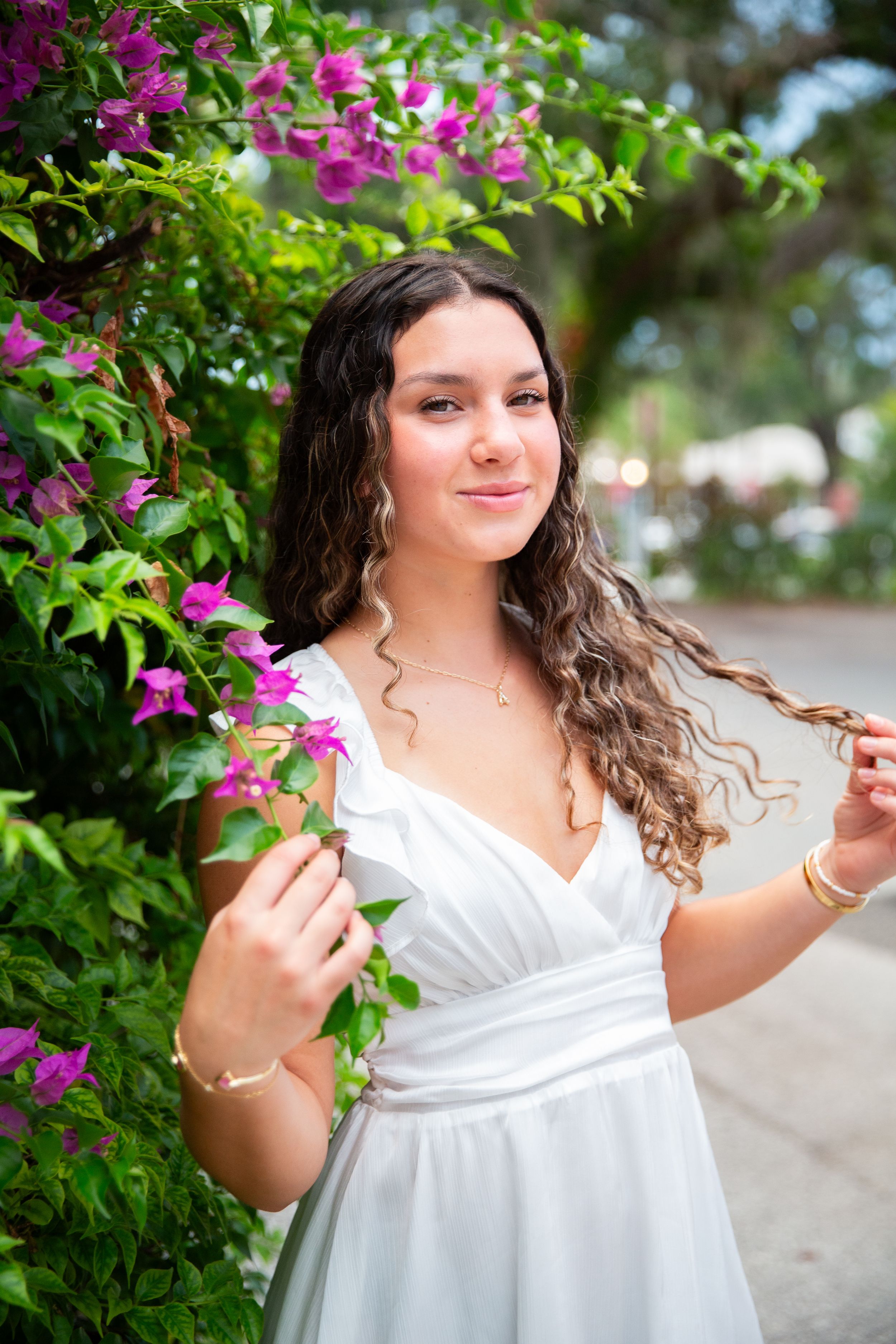 Young woman in white dress poses gracefully beside vibrant pink bougainvillea flowers, twirling her curly hair outdoors.