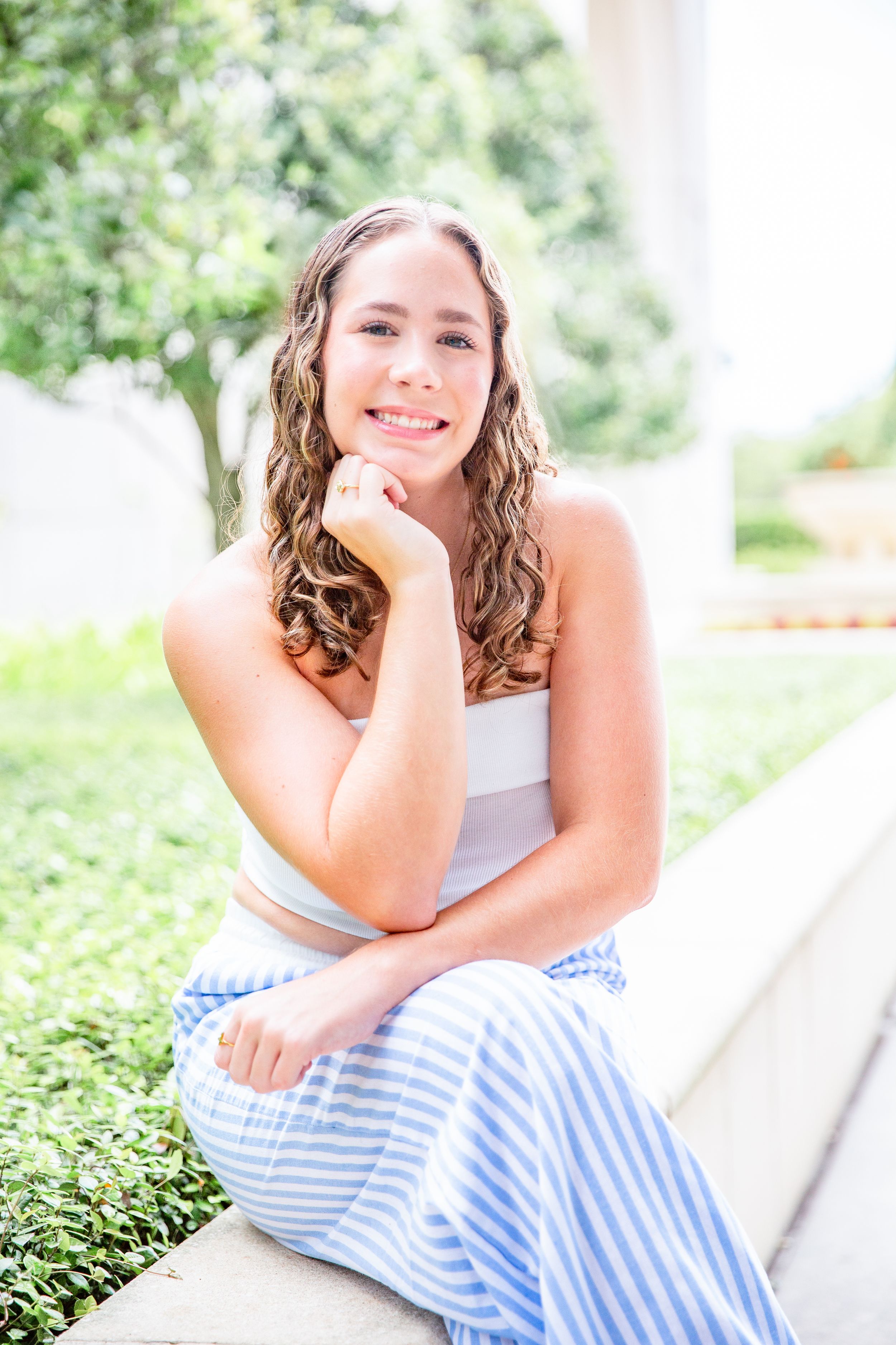 Person in white top and blue striped skirt poses outdoors on white steps surrounded by greenery.