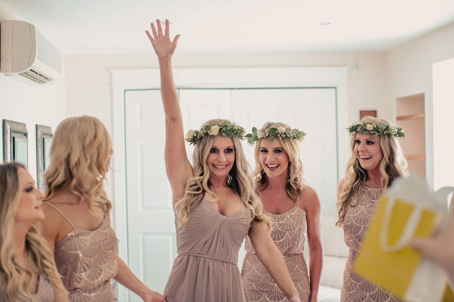 Bridesmaids in blush dresses and floral crowns celebrating together, one raising her hand joyfully before a wedding ceremony.