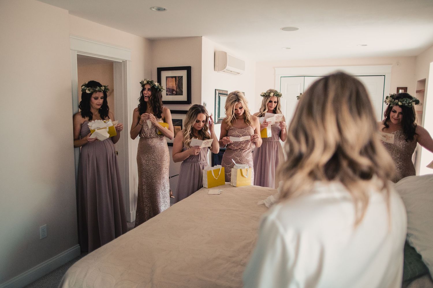 Bridesmaids in dusty rose gowns and floral crowns holding bouquets react to bride in white robe during wedding morning prep.