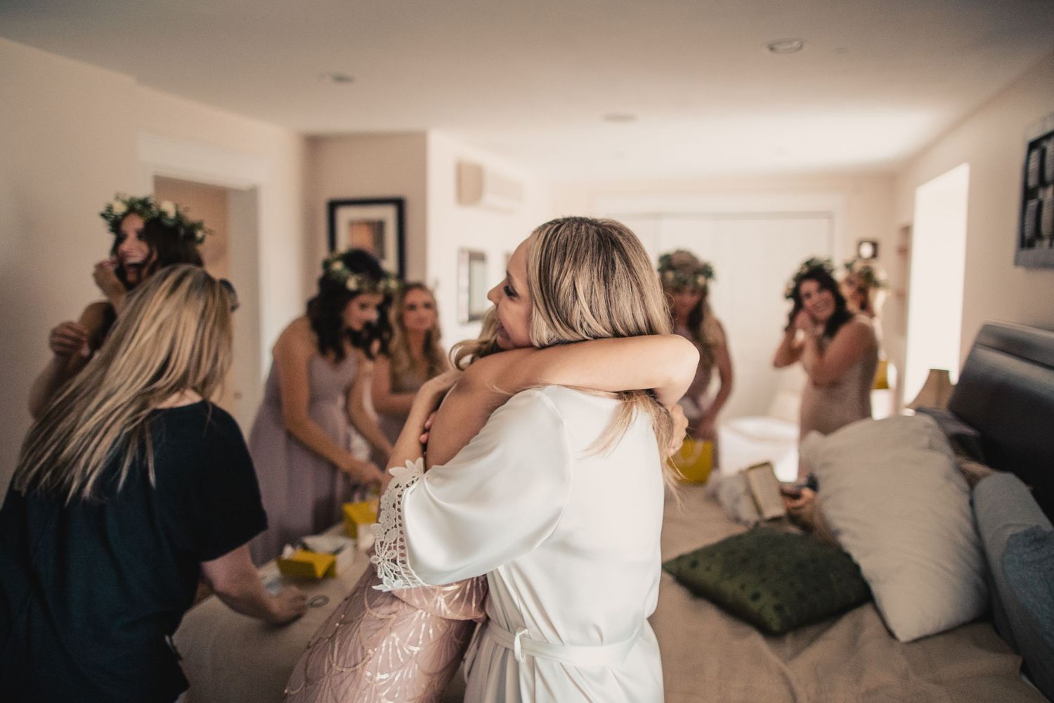 Bride hugging bridesmaid in white robe while bridesmaids in floral crowns gather in bright bedroom.