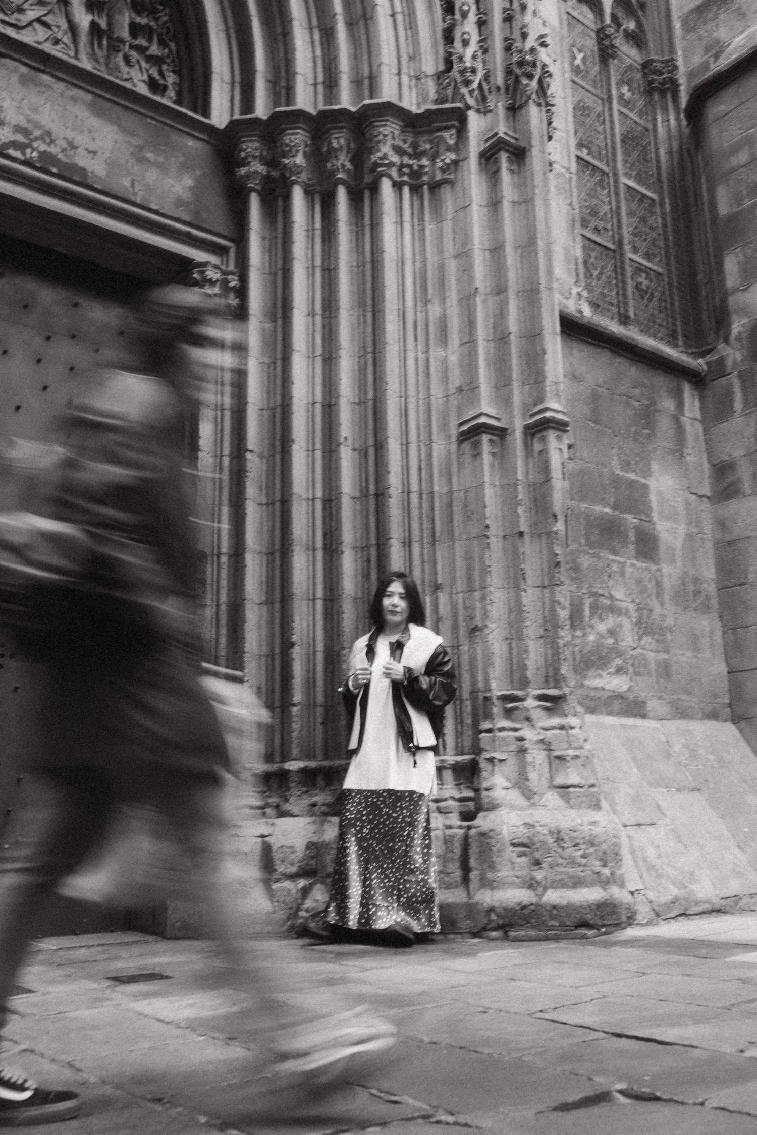 Black and white photo of a woman standing still before a Gothic cathedral as blurred pedestrians pass by.