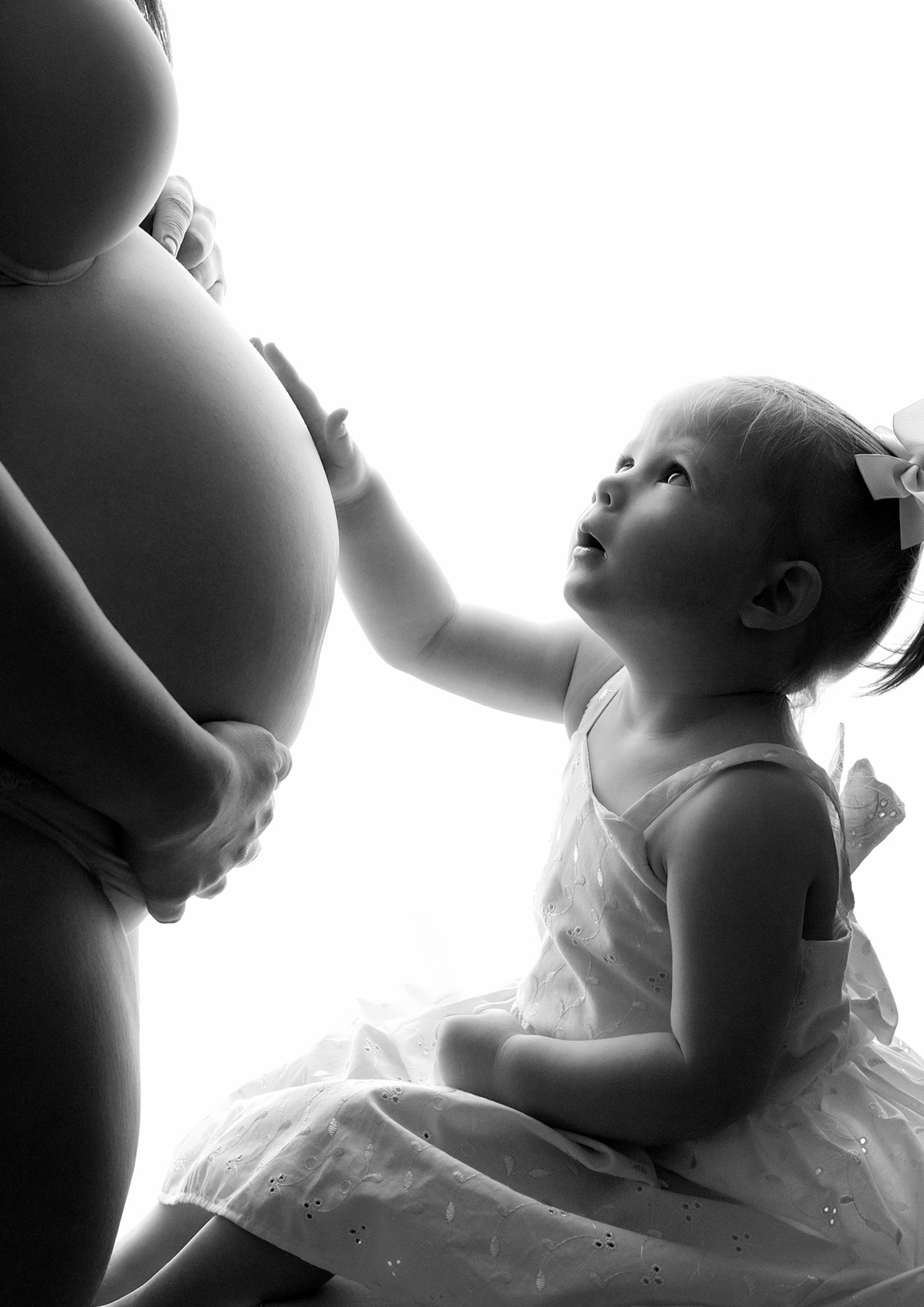 Black and white photo of a toddler girl touching a pregnant woman's belly in a tender maternity portrait.