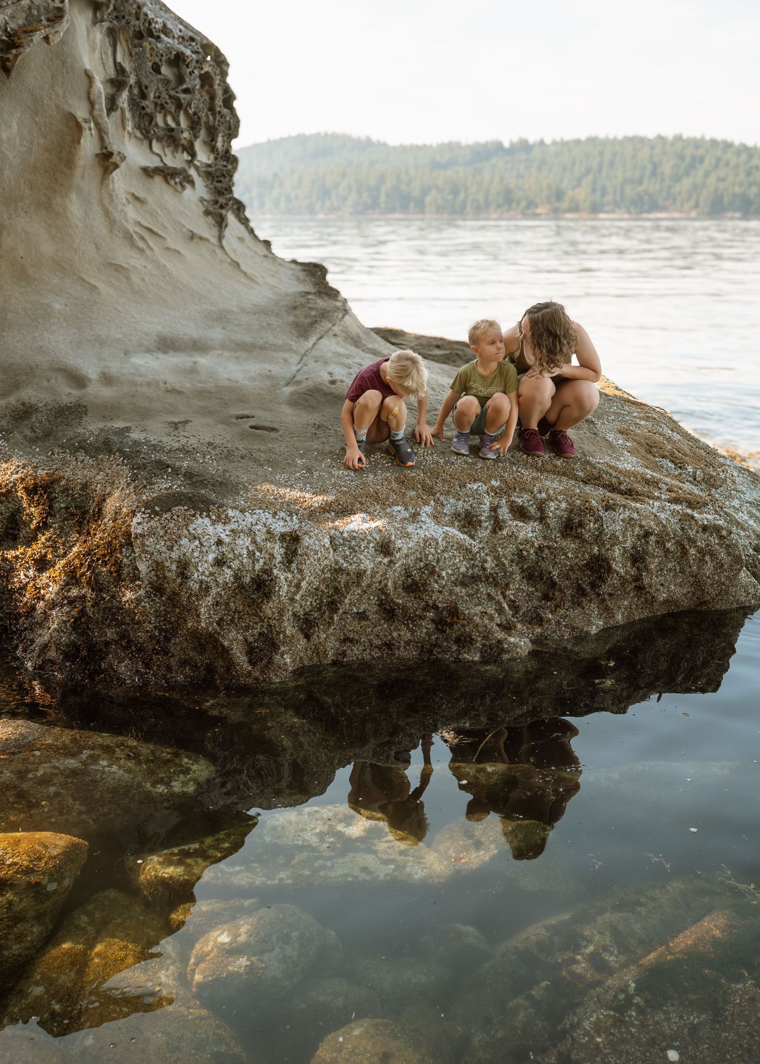 Three children explore tide pools on a rocky shoreline with calm water and forested hills in the background.