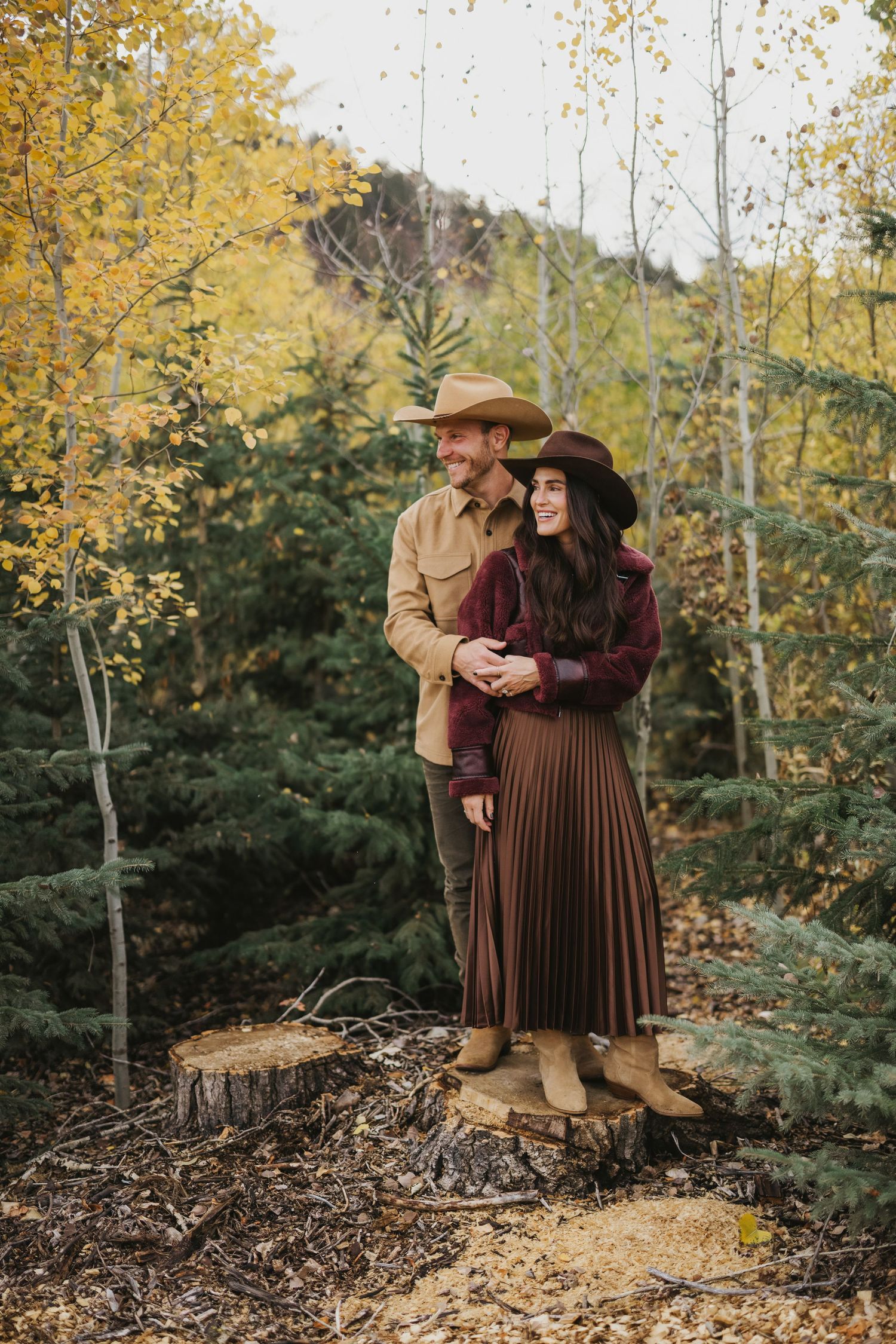 A couple in western attire share a tender moment outdoors during autumn with yellow foliage in the background.