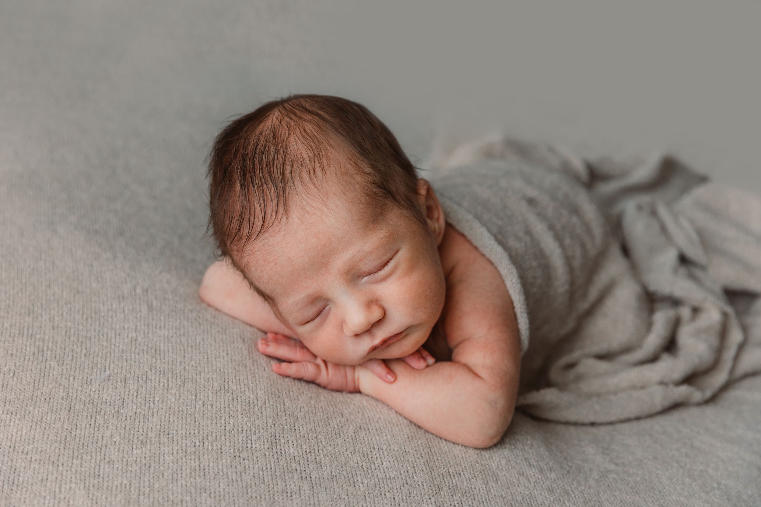 Sleeping newborn baby wrapped in grey blanket on neutral background.