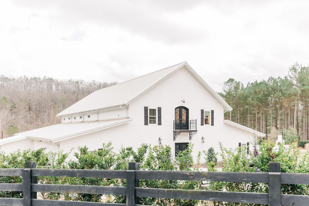 White farmhouse style home with black fencing and well-manicured landscaping sits on a sunny rural property.