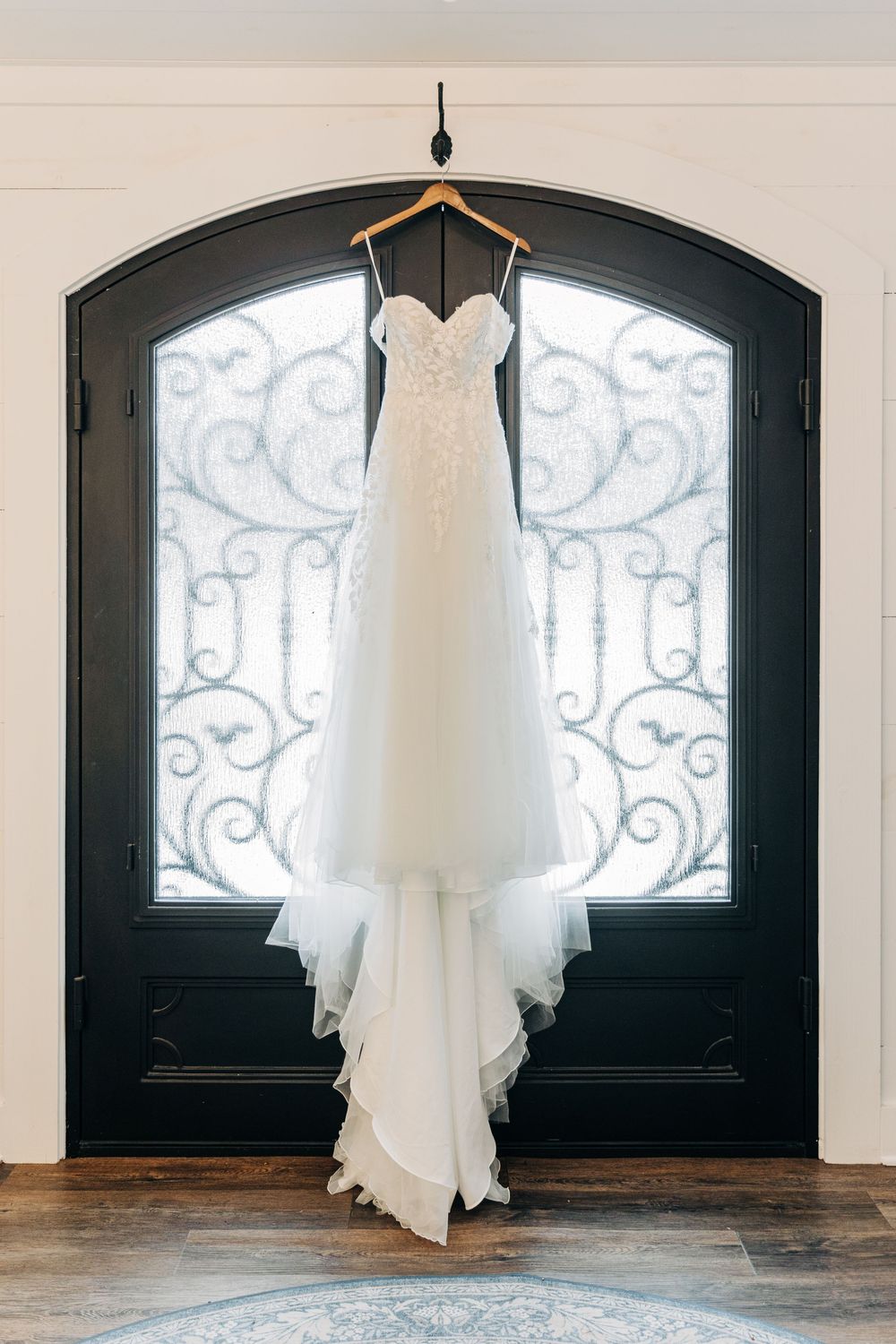 A white wedding dress hangs elegantly in front of an arched window with decorative iron scrollwork.