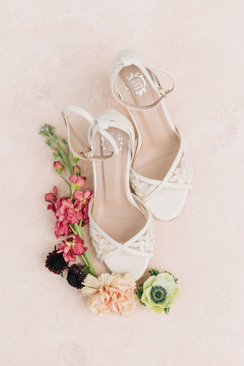 White bridal heels surrounded by delicate pink and burgundy flowers on a soft beige background.