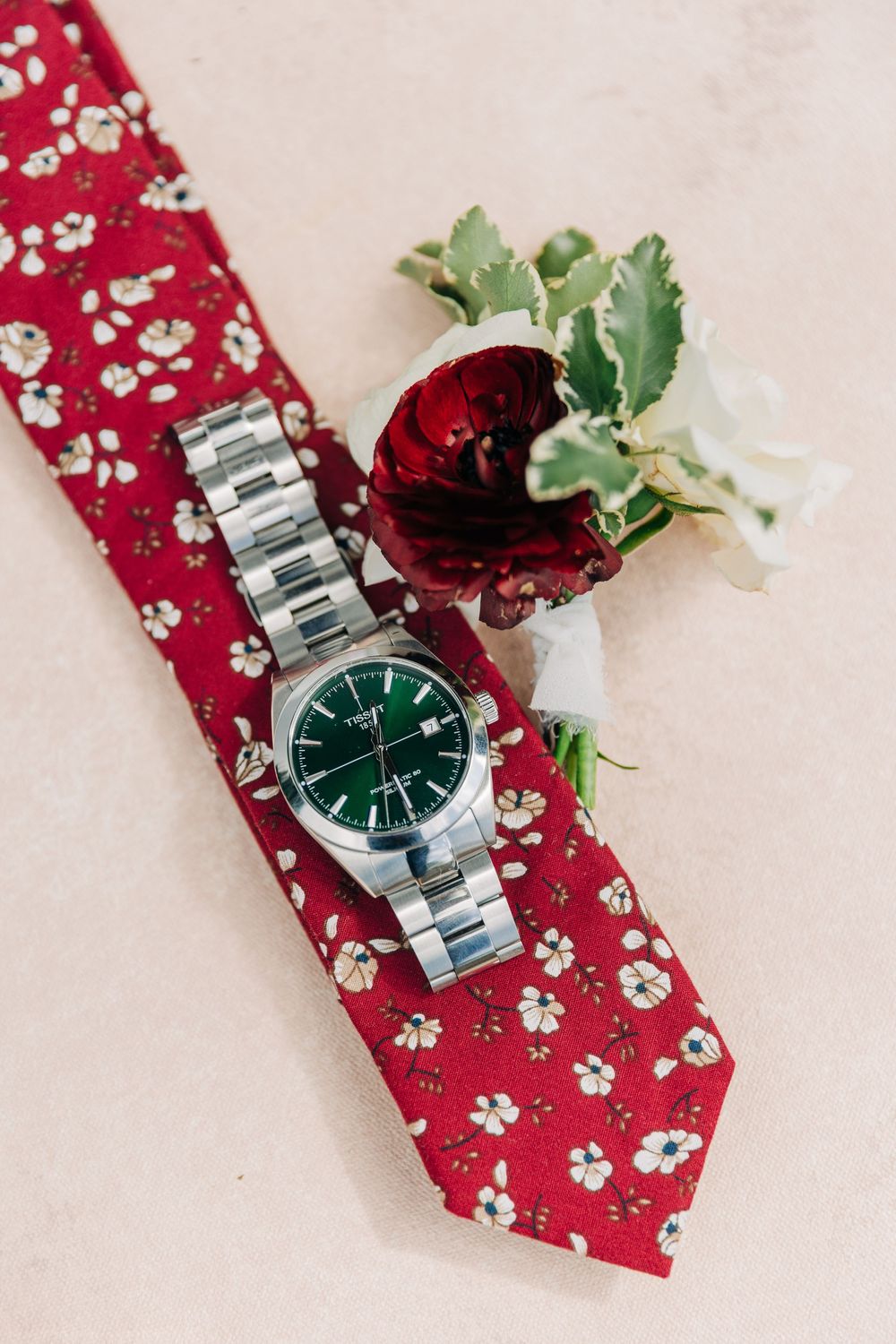A silver wristwatch with green dial face displayed on a red floral patterned tie with decorative flowers.