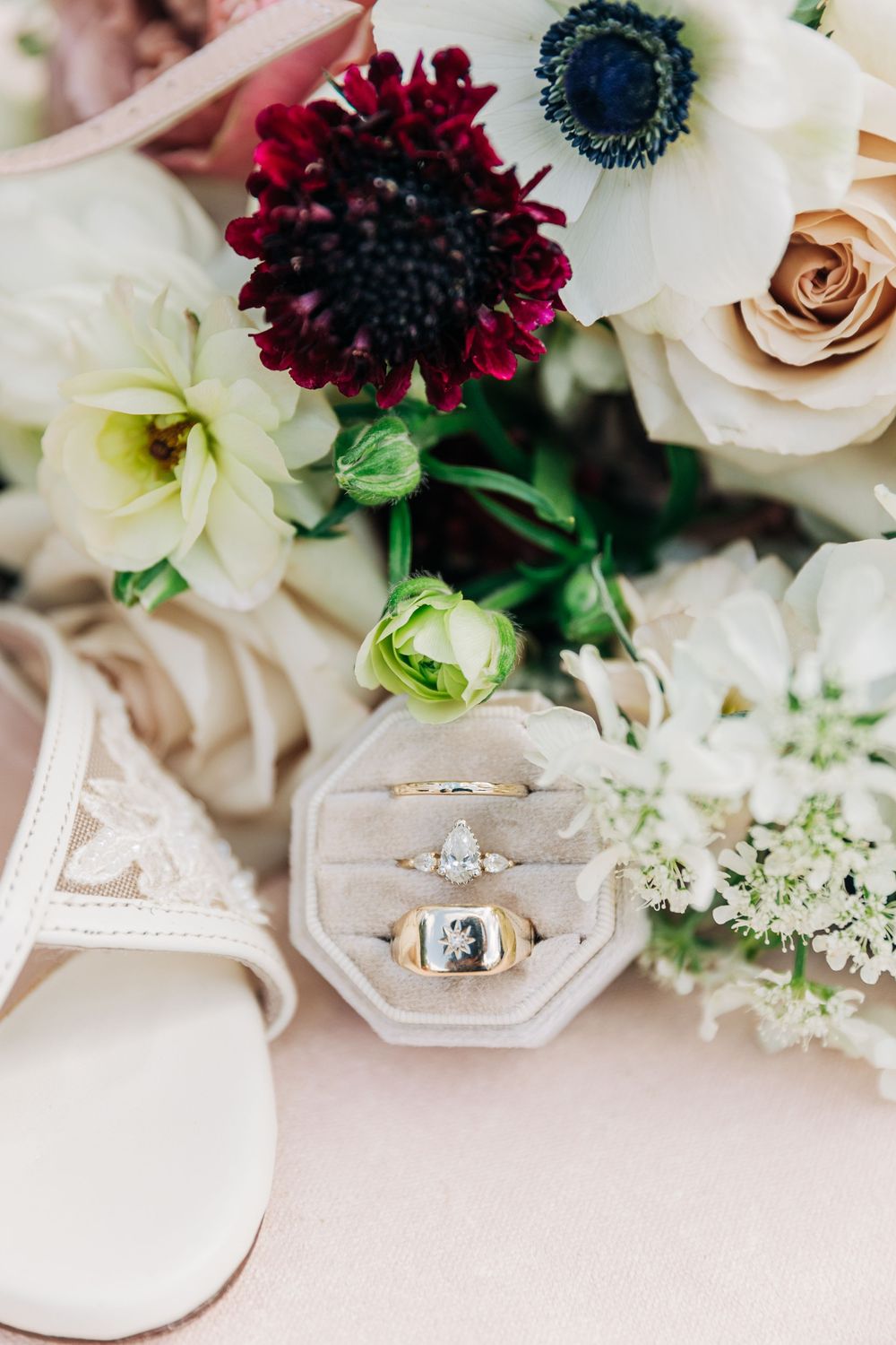 Wedding rings and engagement rings displayed with dark burgundy and white flowers on a light beige surface.
