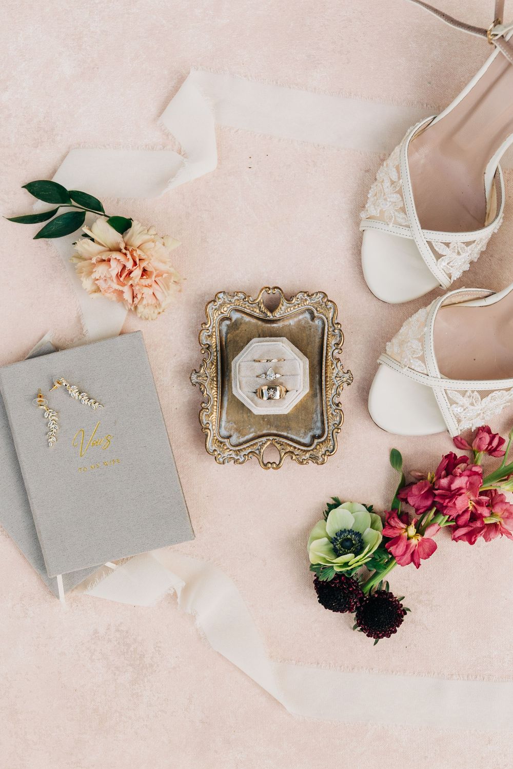 Wedding accessories including white shoes, vintage frames, and floral decorations arranged on a blush background.