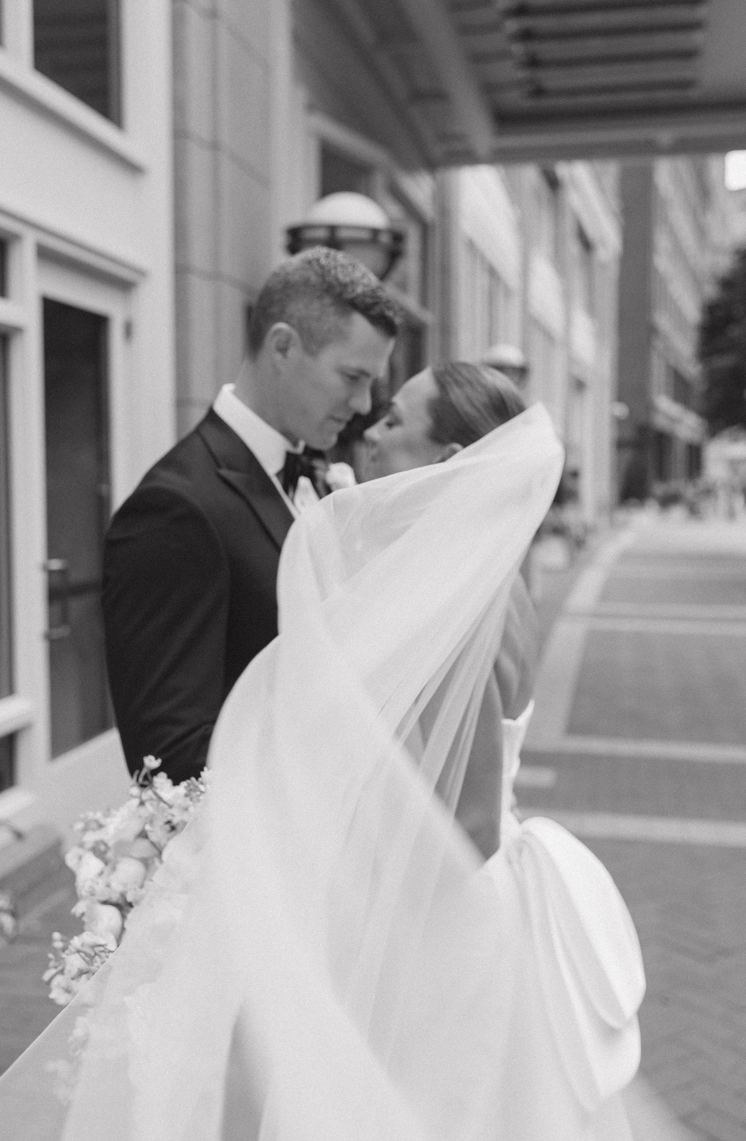 A romantic black and white wedding photo showing a tender moment between newlyweds on a city sidewalk.
