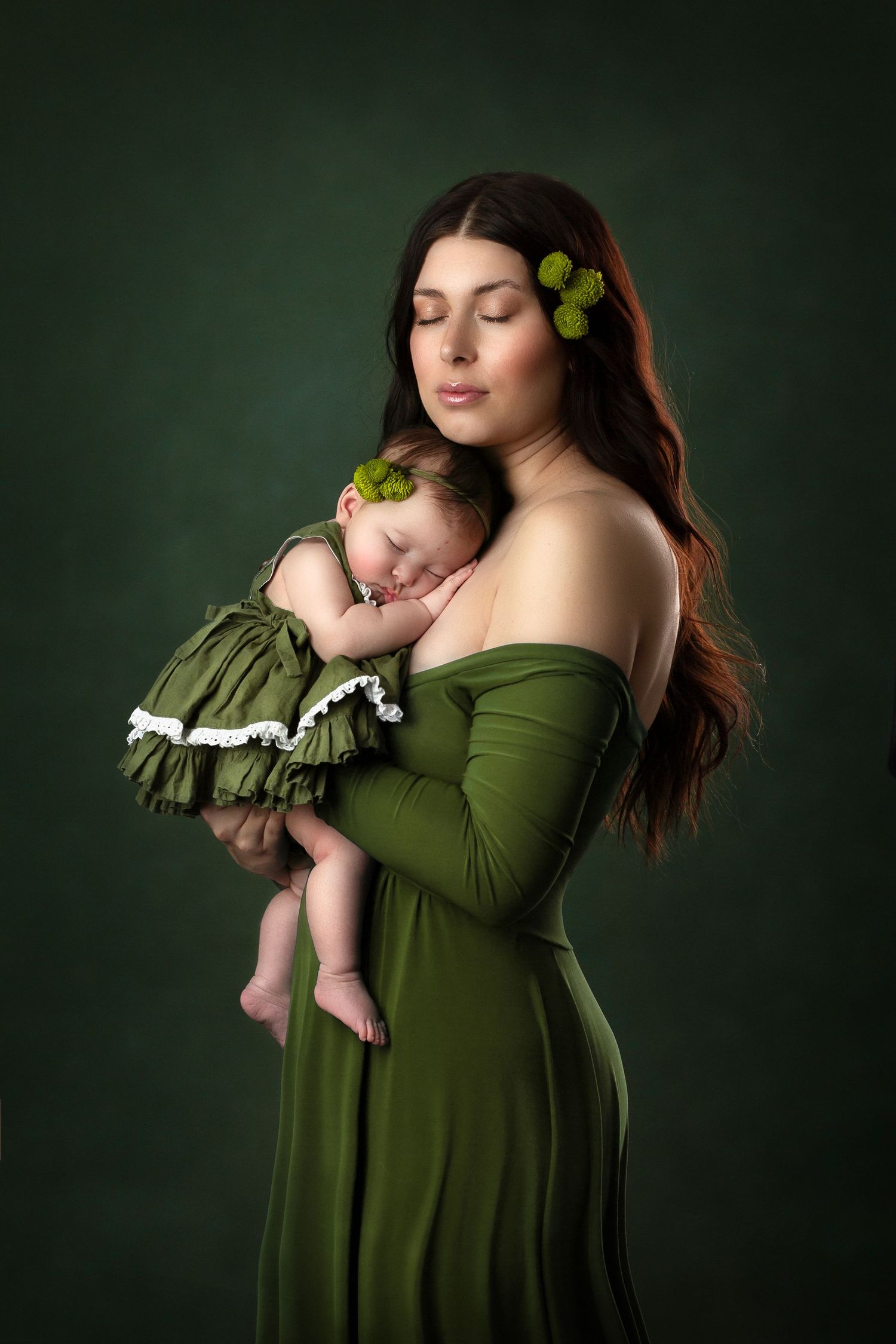 A tender portrait in green tones shows a mother and sleeping baby in matching emerald dresses against a dark background.