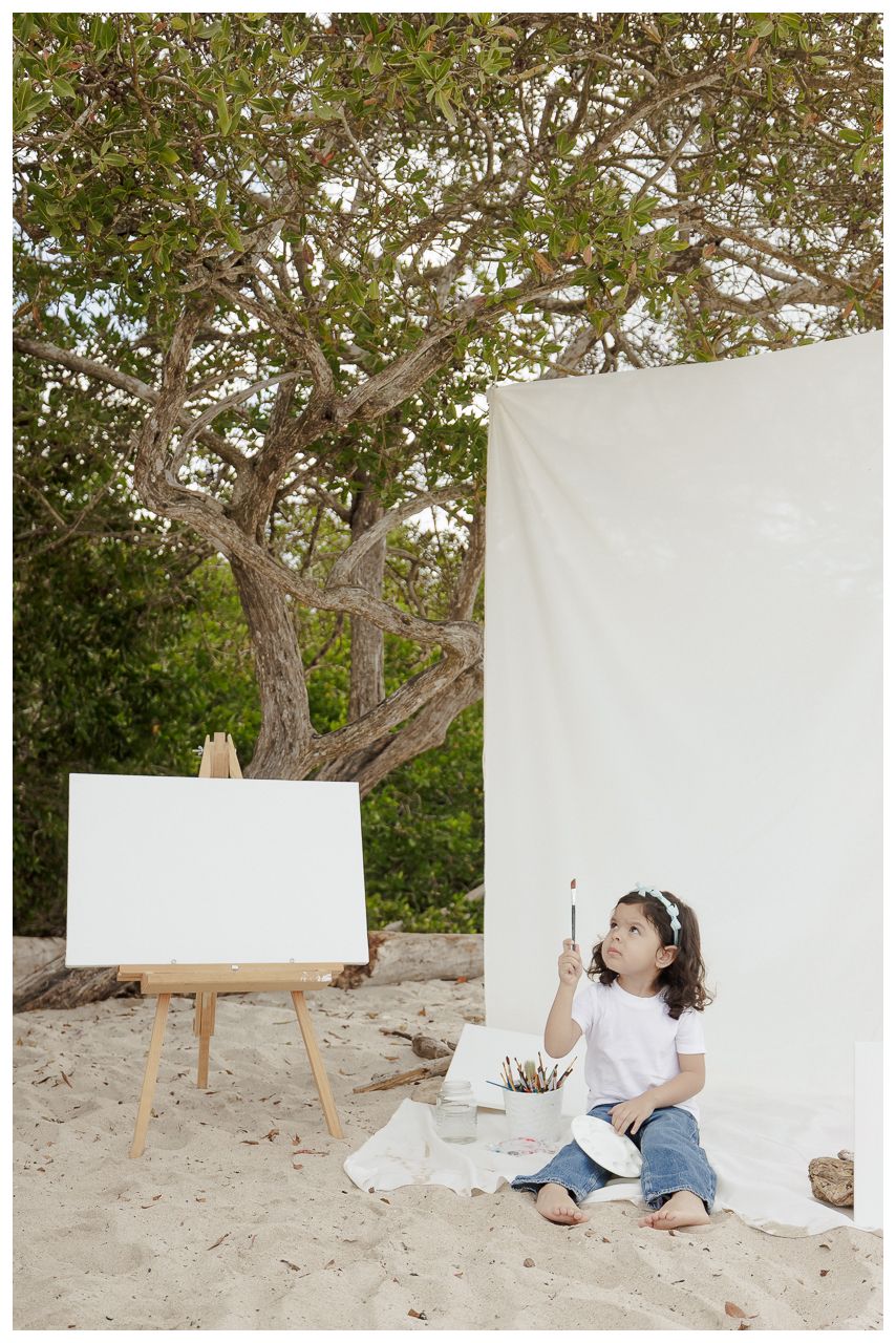 A series of white canvases set up on easels along a sandy beach with trees in the background.