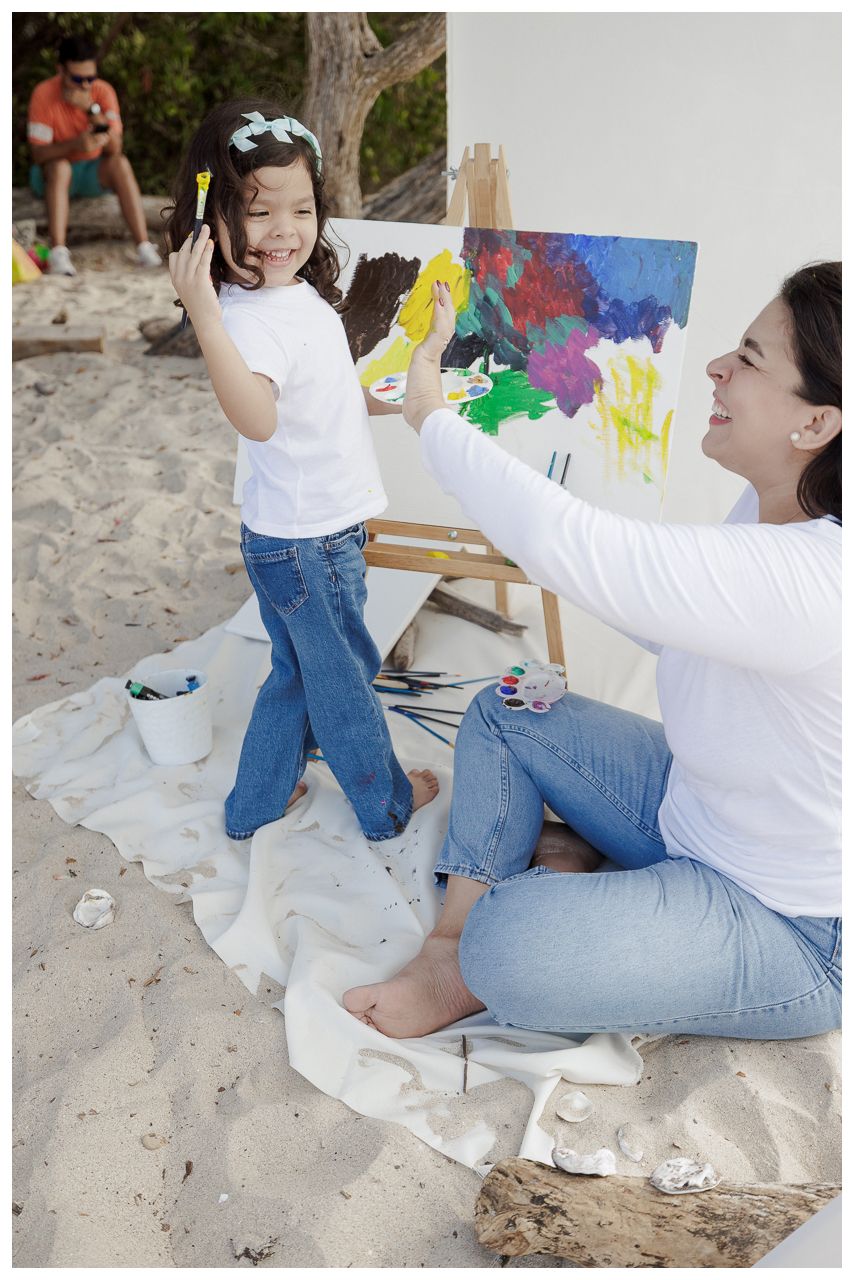 A parent and child painting together outdoors on a sunny day, sitting on a white sheet on the ground.