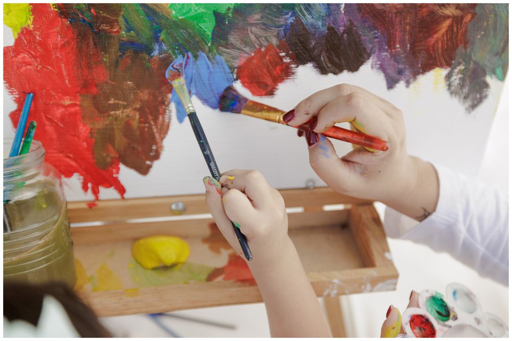 Child's hands holding colorful art supplies and painting materials on a white surface.