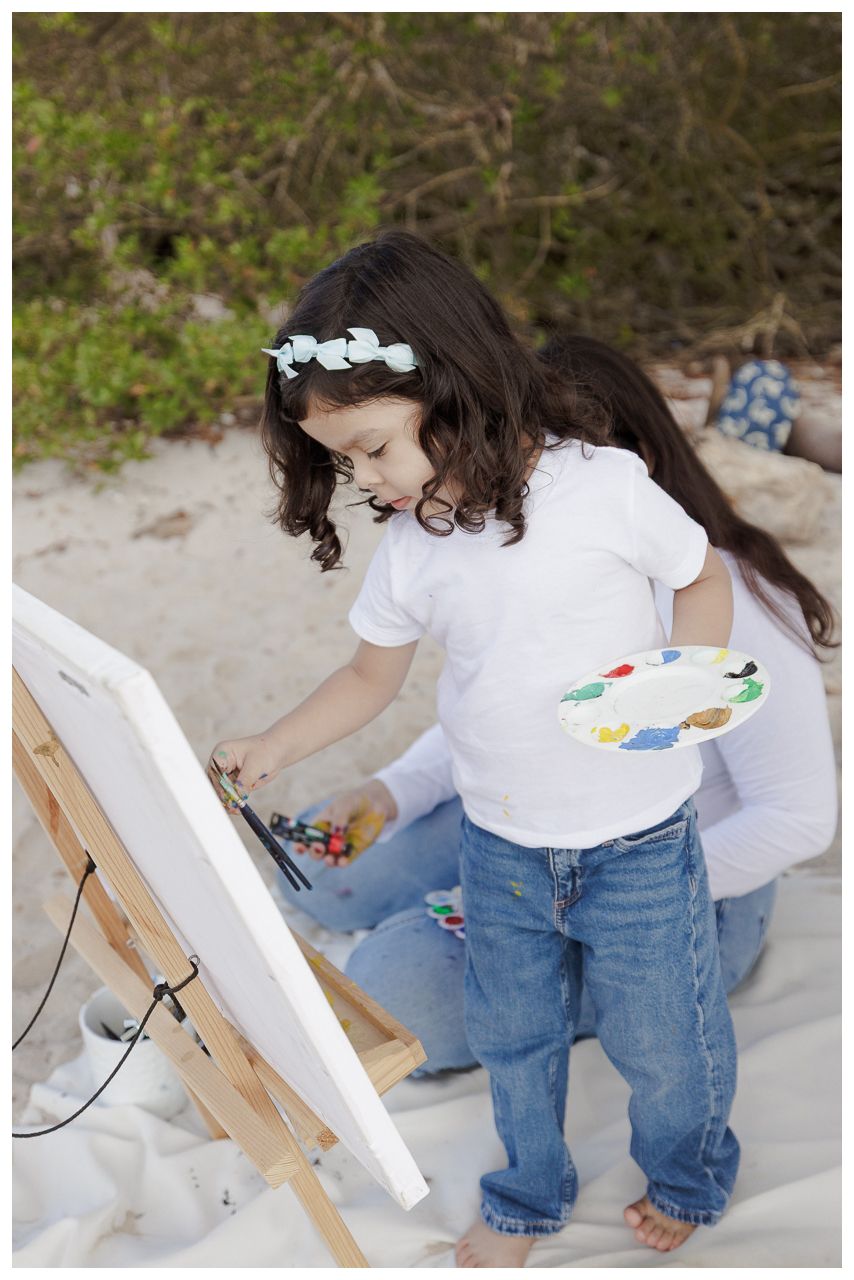 A child in a white shirt and jeans sits on a sandy beach while painting on a wooden easel.