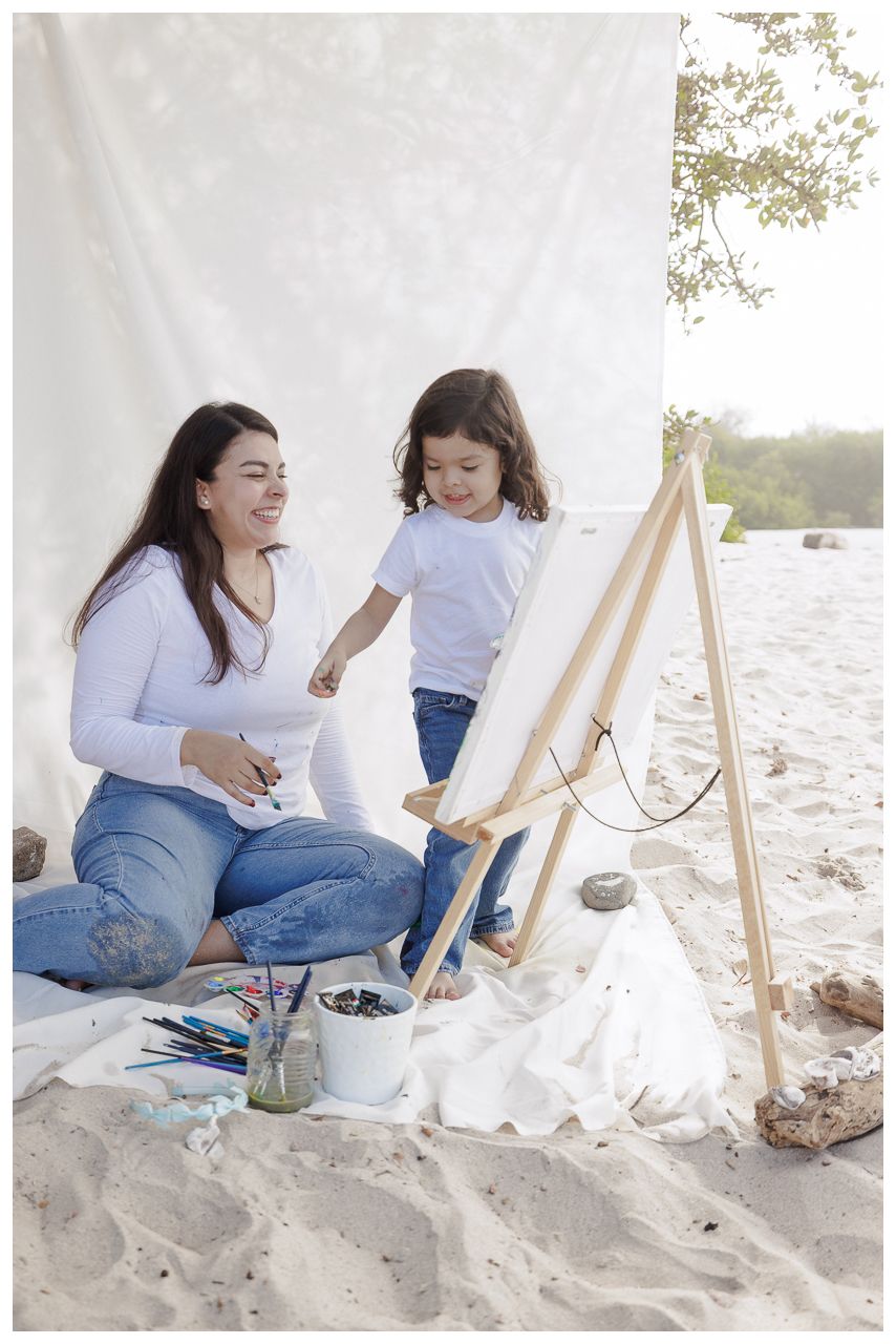 A parent and child sit on a sandy beach with an art easel, enjoying a creative painting activity together.