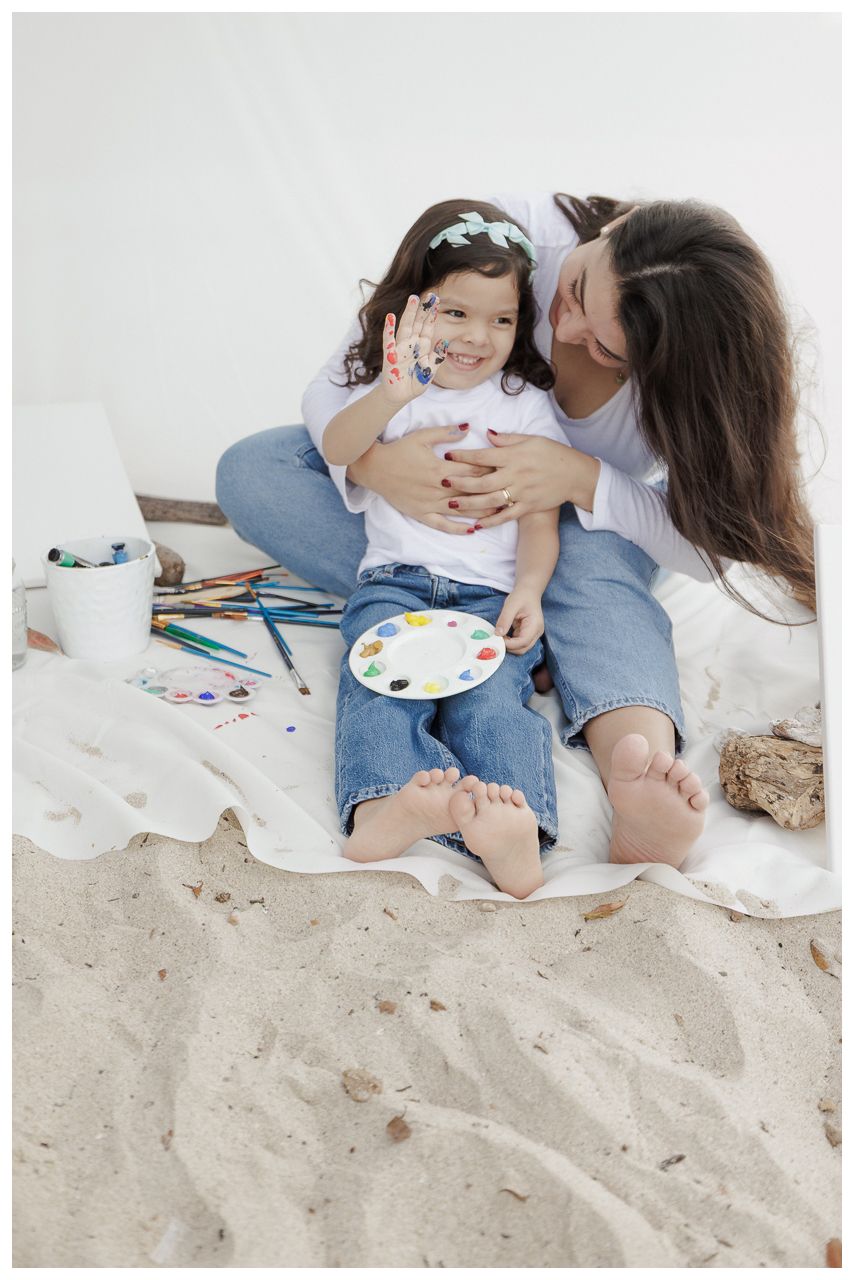 Two people sit together on a sandy beach wearing denim while sharing a painting palette and creating art.