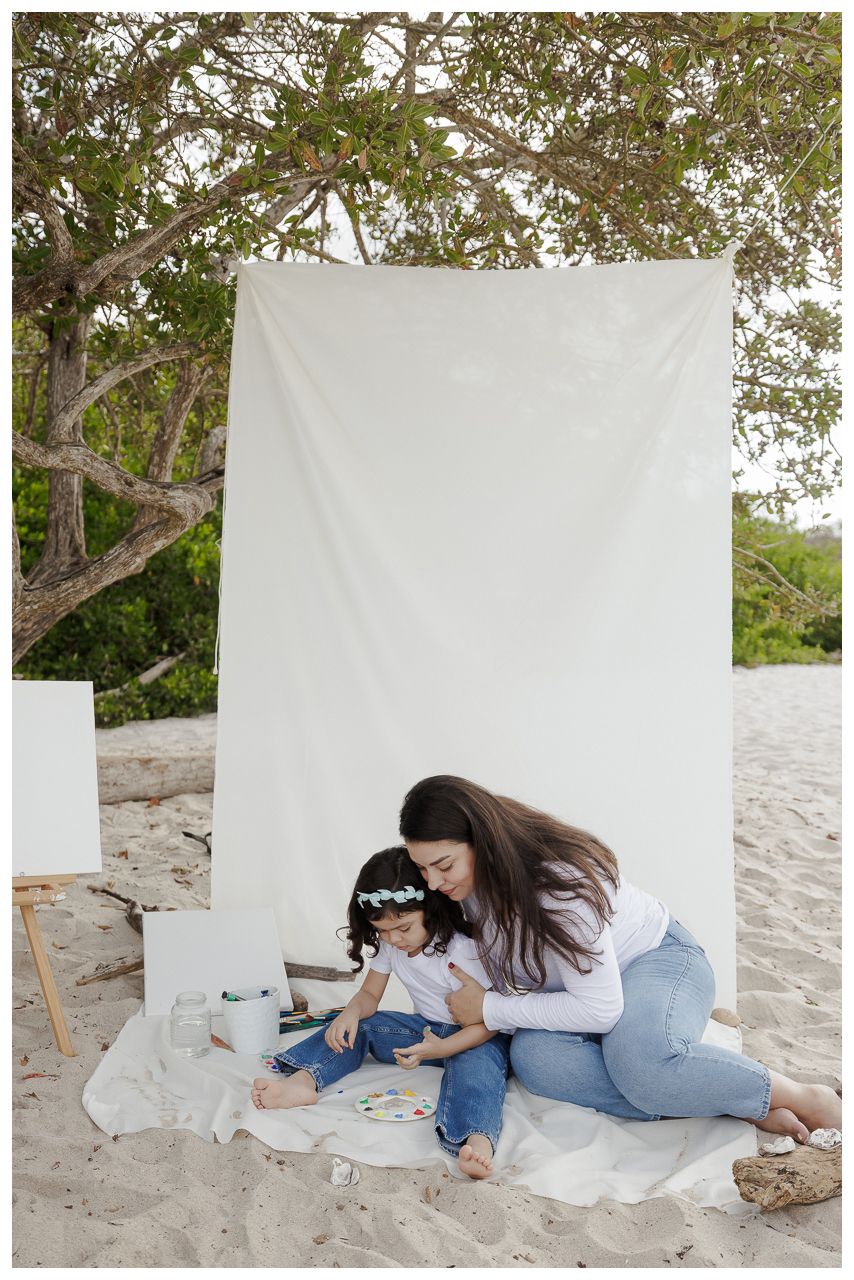 A parent and child sit together on a sandy beach against a white backdrop during a photo session.