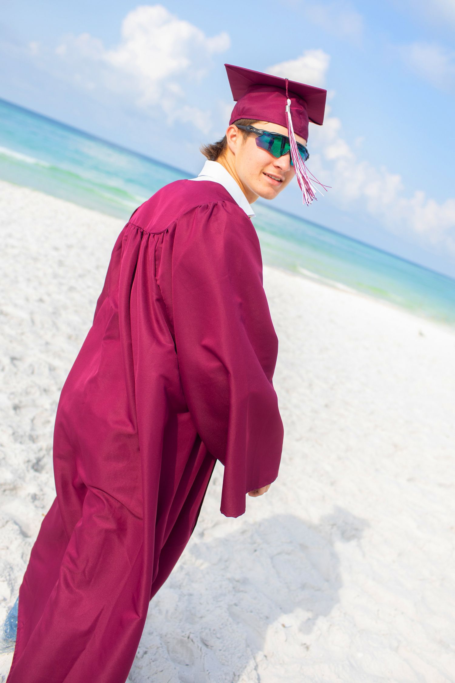 Graduate in burgundy cap and gown poses on a sunny beach with turquoise ocean waters and white sand.