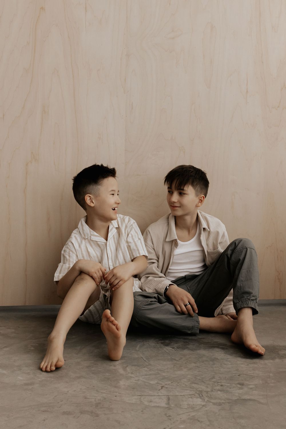 Two children in casual white and gray clothing sit together on a concrete floor in a minimalist setting.
