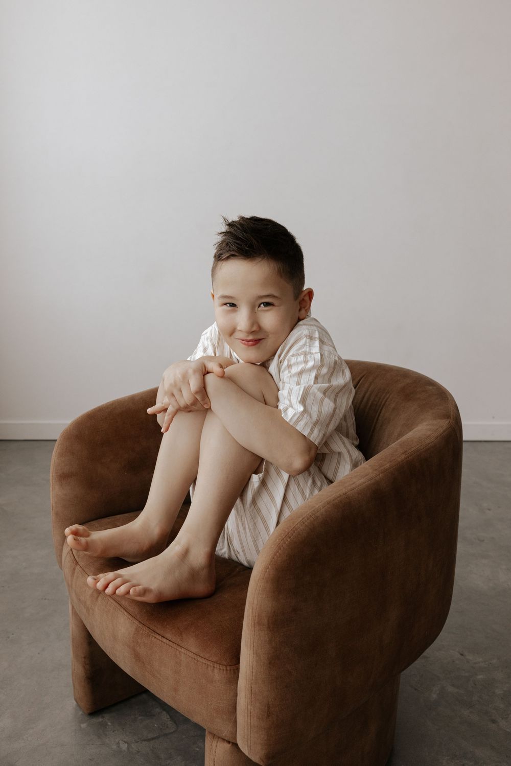 A small child in white clothing sits playfully on a brown velvet armchair against a light gray background.