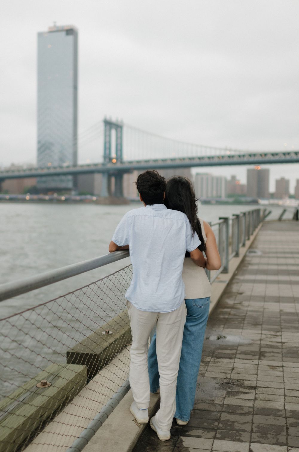 Couple walking together along a waterfront promenade with city skyline and bridge in the background.