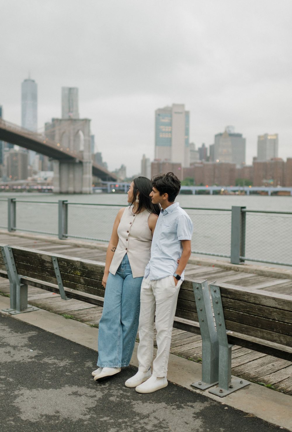 A couple sits together on a wooden pier overlooking a cityscape on a cloudy day.