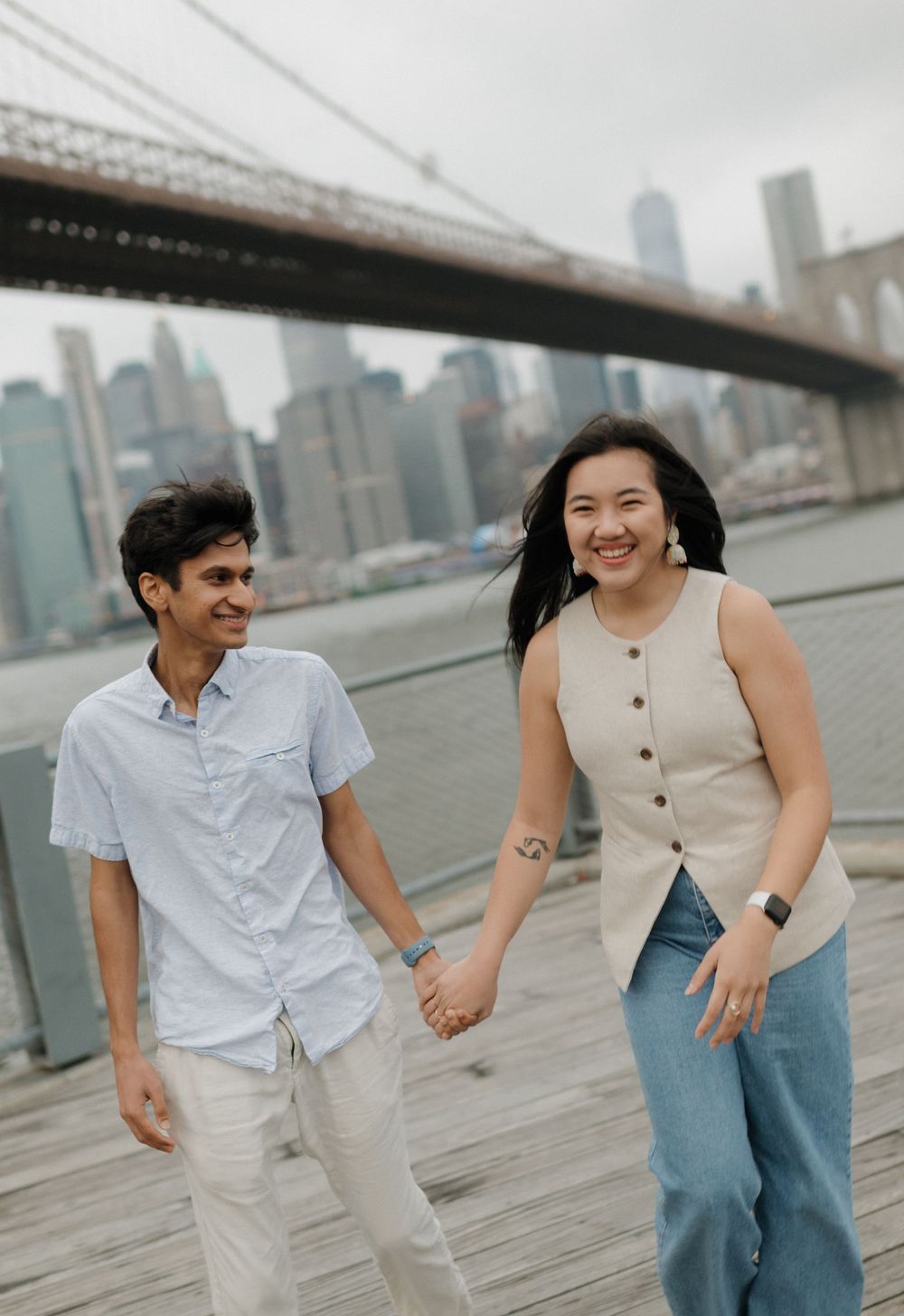 A couple holds hands while walking along the waterfront near the Brooklyn Bridge with the NYC skyline in the background.