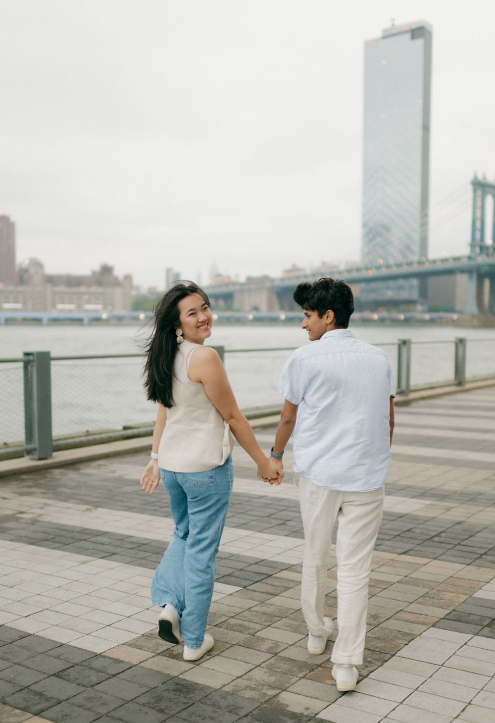 A couple in white and blue outfits holds hands while dancing playfully along a waterfront promenade with city buildings behind them.