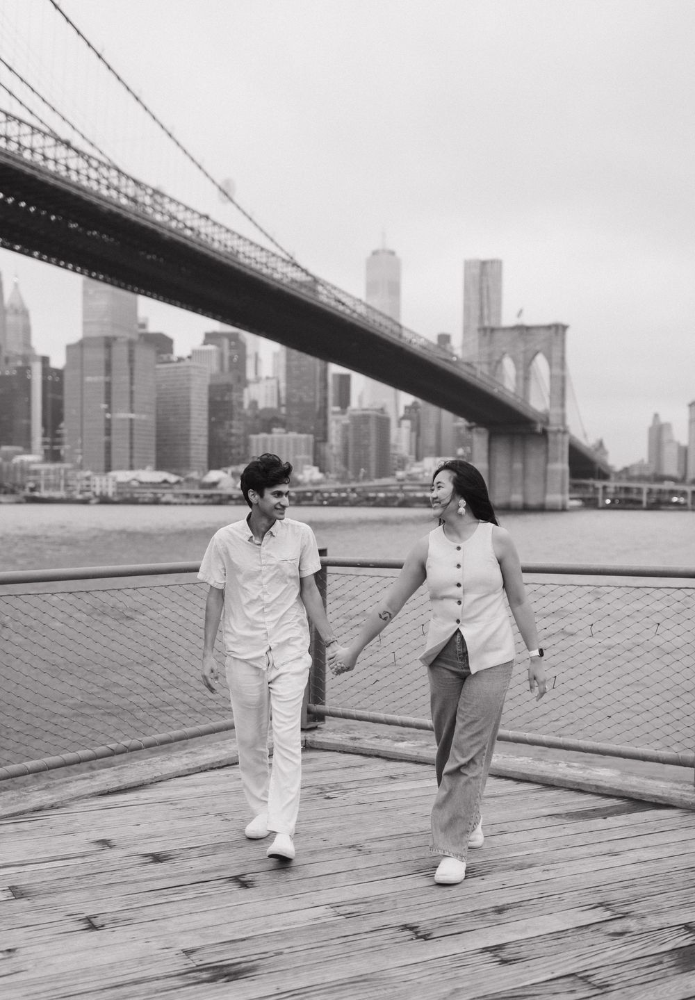 Two people in white and dark clothing sit and converse on a wooden pier with the Brooklyn Bridge visible in background.