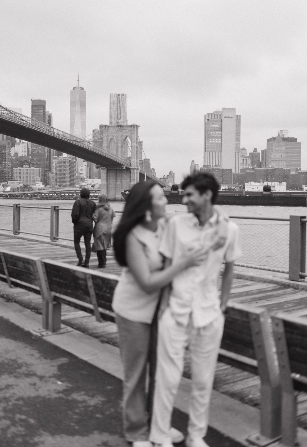A couple embraces playfully on a waterfront walkway with urban buildings in the background.