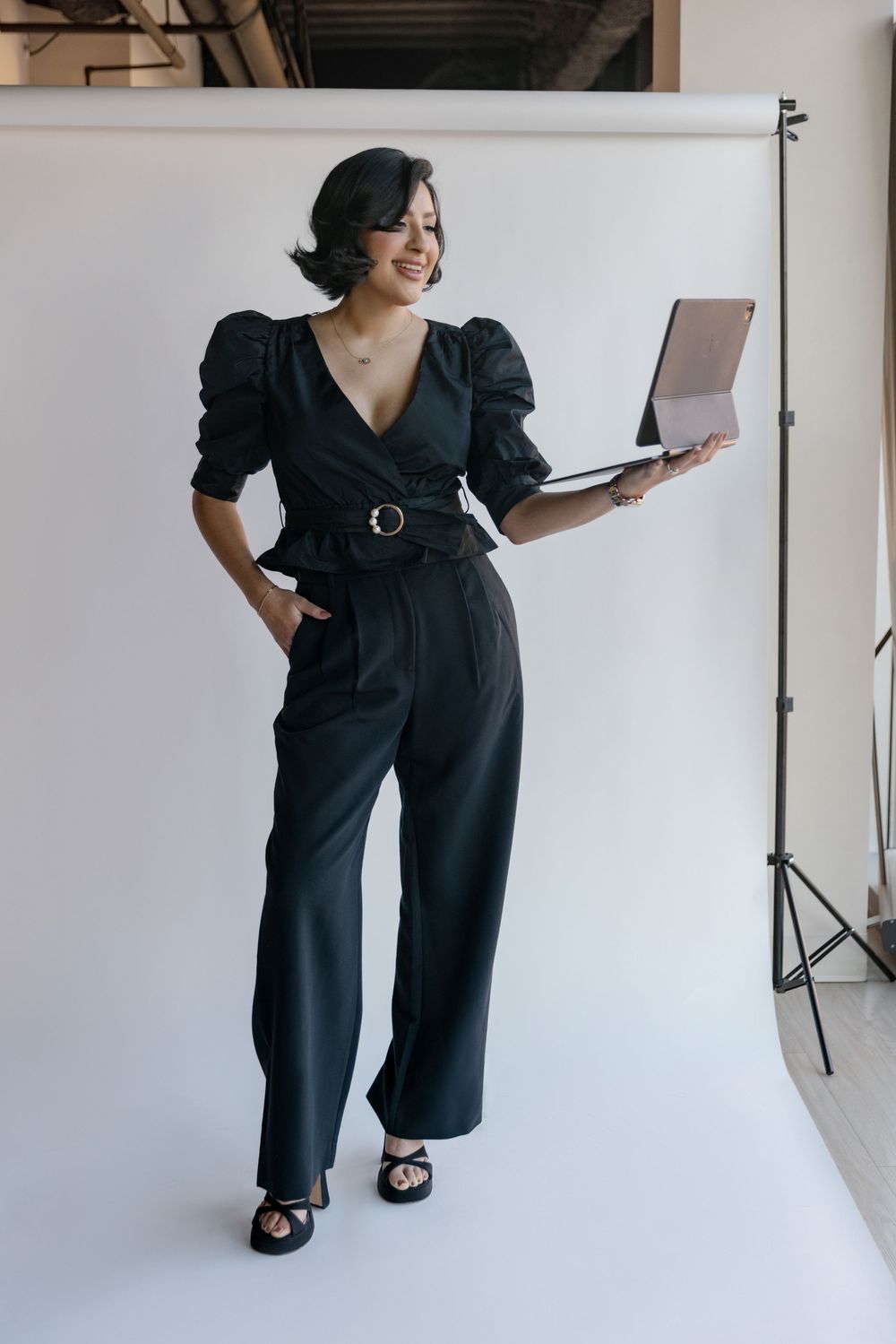 Person in black puff sleeve jumpsuit with belt detail poses against white backdrop in studio photoshoot.