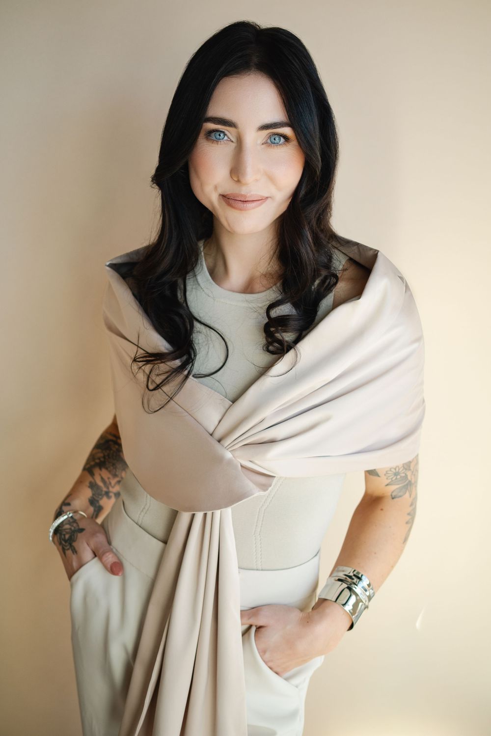 Portrait of someone in a cream-colored wrap dress with dark hair and tattoos against a neutral background.