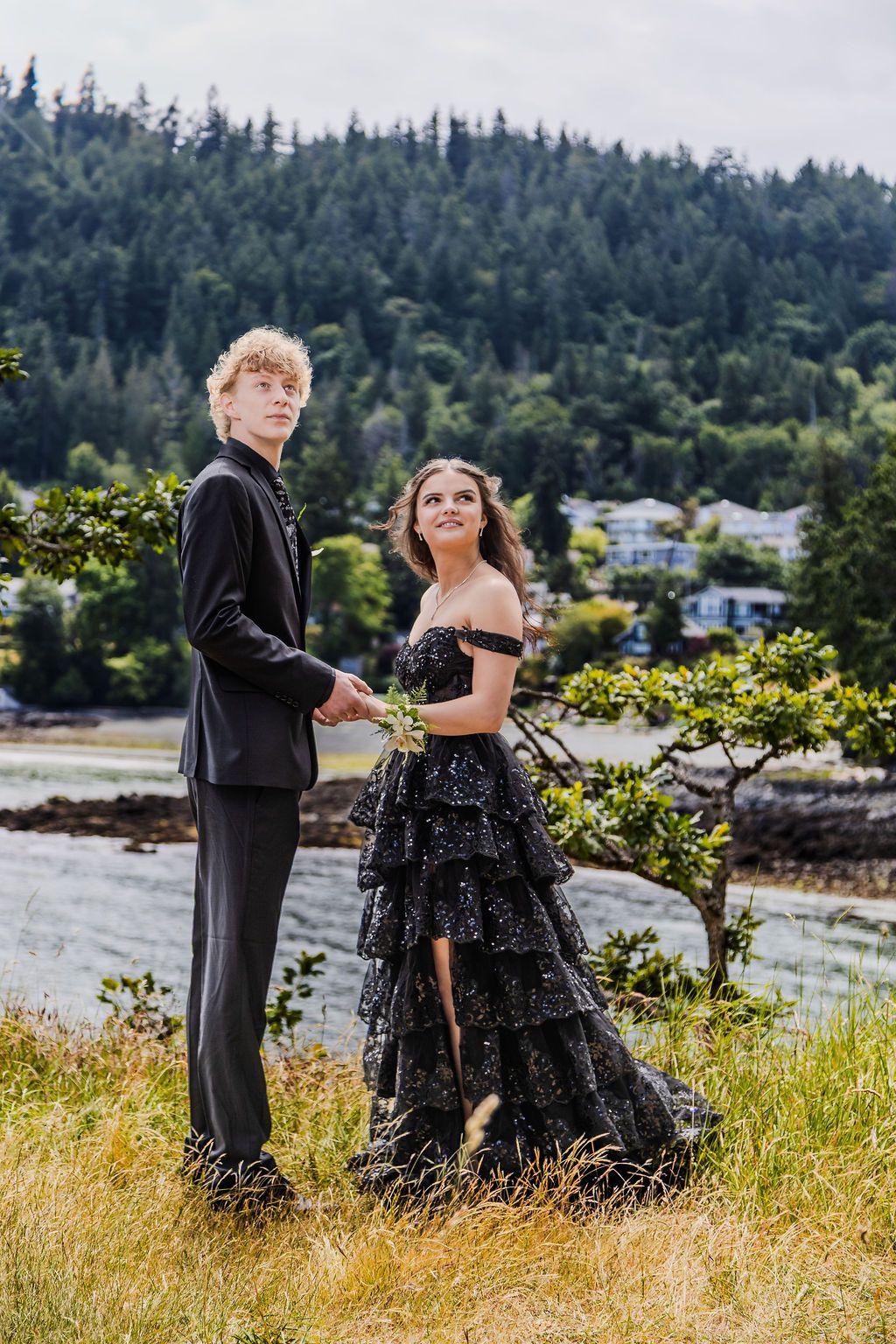 Elegant prom couple in black formal wear hold hands outdoors with a forested waterfront behind them.