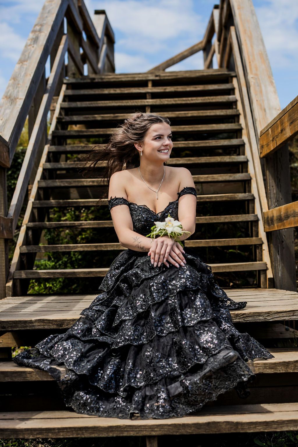 Smiling teen in black tiered sequin prom gown sitting on wooden stairs holding white bouquet.